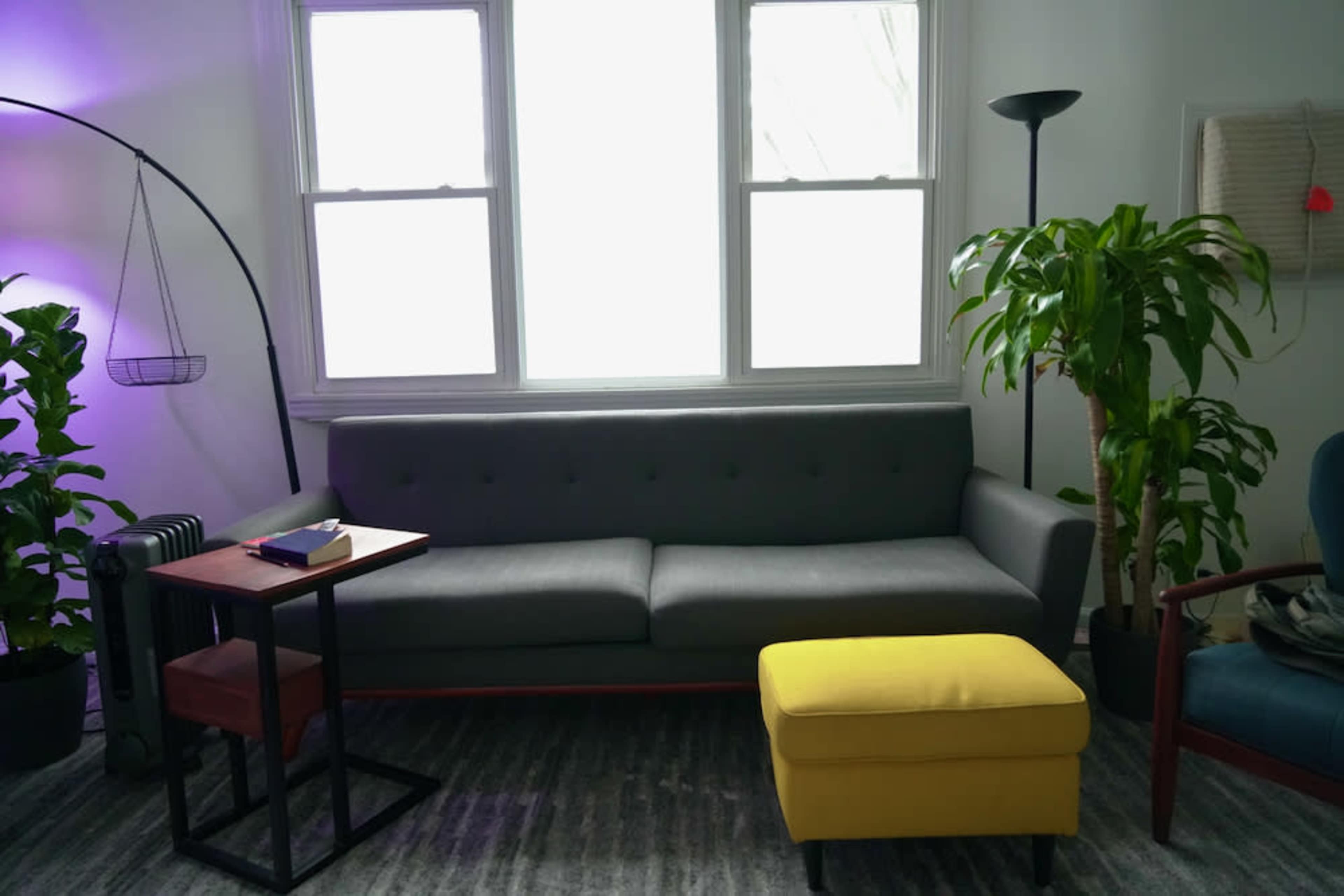 A living room with a gray couch, a small wooden side table, a yellow ottoman, and several plants near the windows.