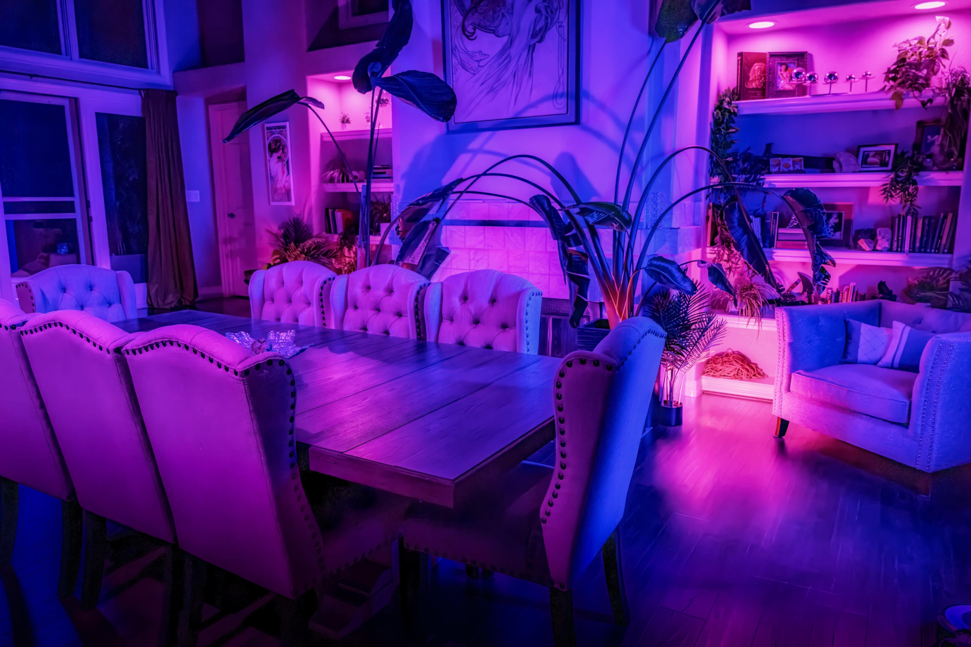 A modern dining area features a large wooden table surrounded by upholstered chairs, illuminated by vibrant purple lighting and accented with decorative plants.