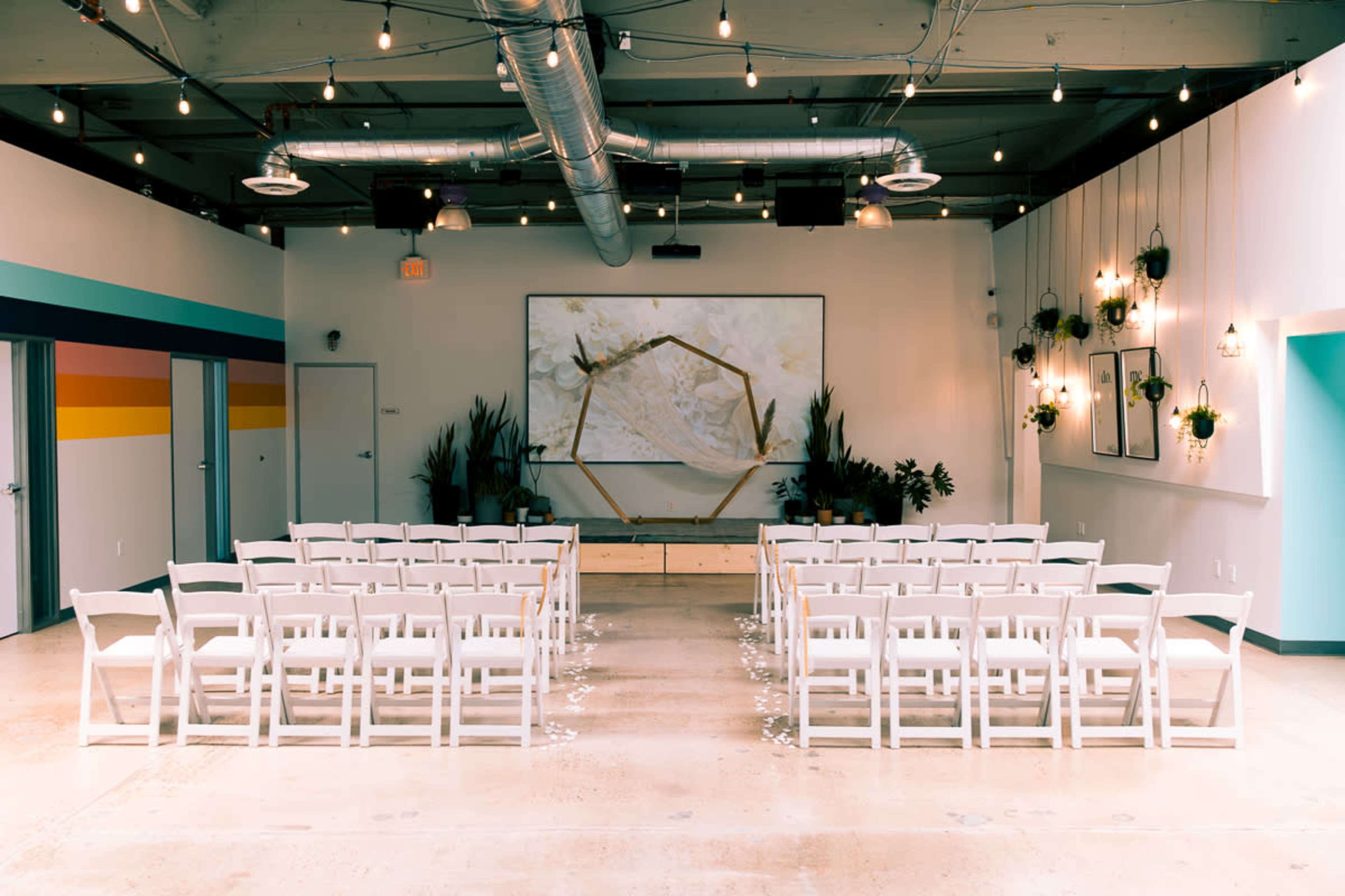 The image shows a spacious event room set up with white folding chairs facing a large screen, decorated with plants and string lights overhead.