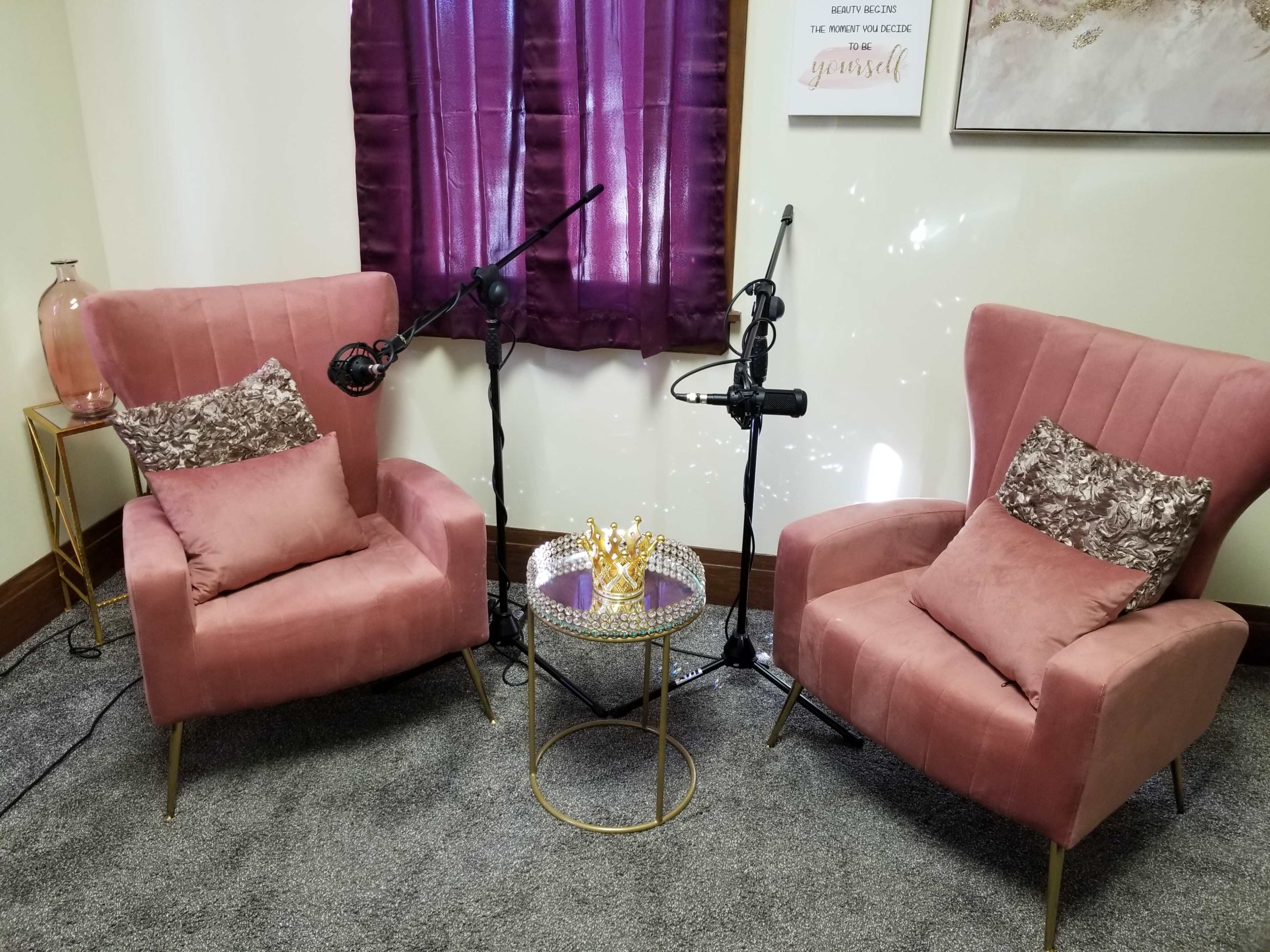 Two pink armchairs with decorative pillows placed next to a small round table with a crown centerpiece, and microphones positioned nearby for recording.