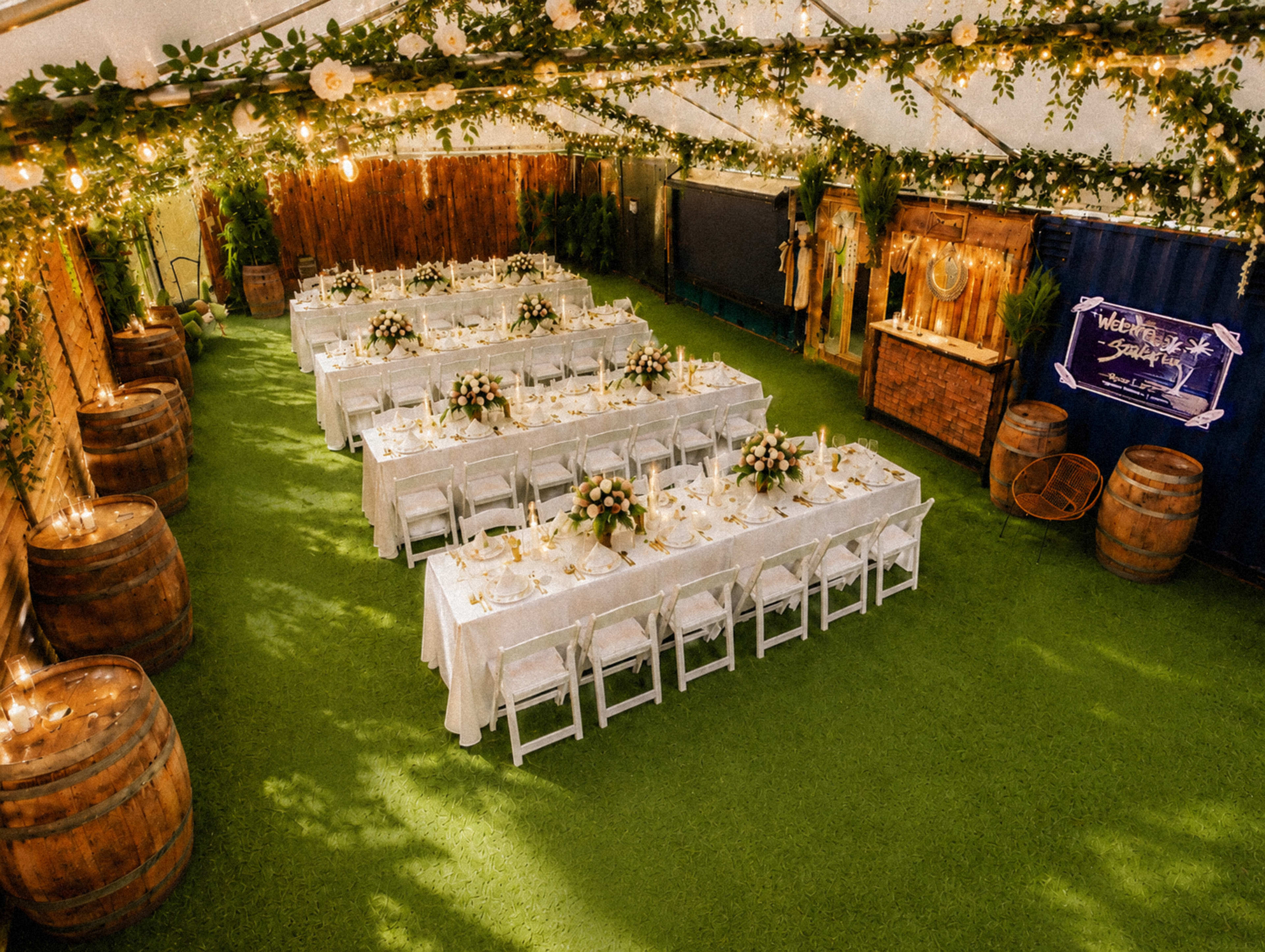 A large tented space features rows of elegantly set tables adorned with flowers, surrounded by greenery and illuminated by string lights.