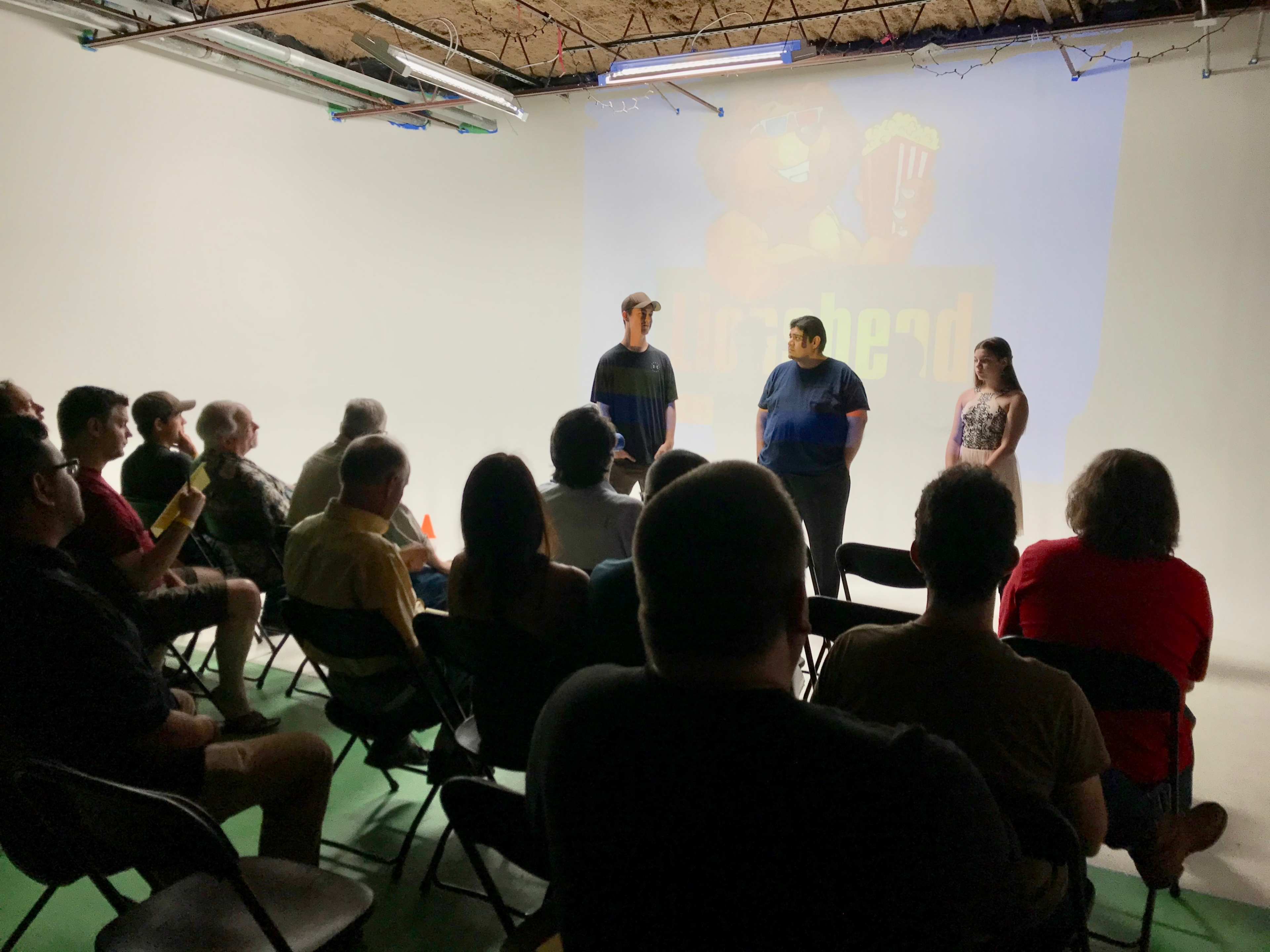 A group of people sits in chairs while three individuals stand in front of a large projector screen displaying an animated character and text.