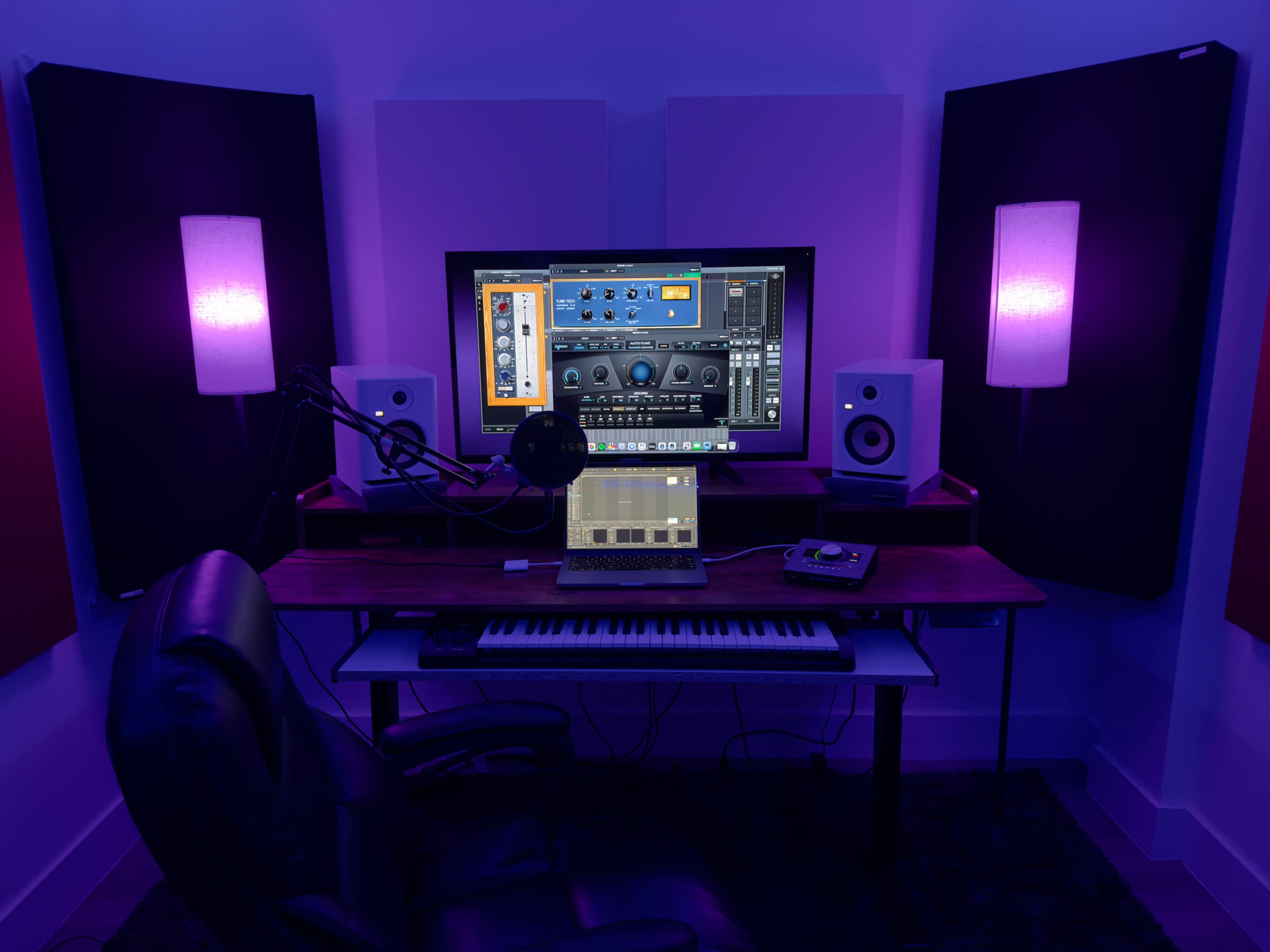 A music production studio features a desk with a computer, audio interface, and keyboard, surrounded by soundproofing panels and purple ambient lighting.