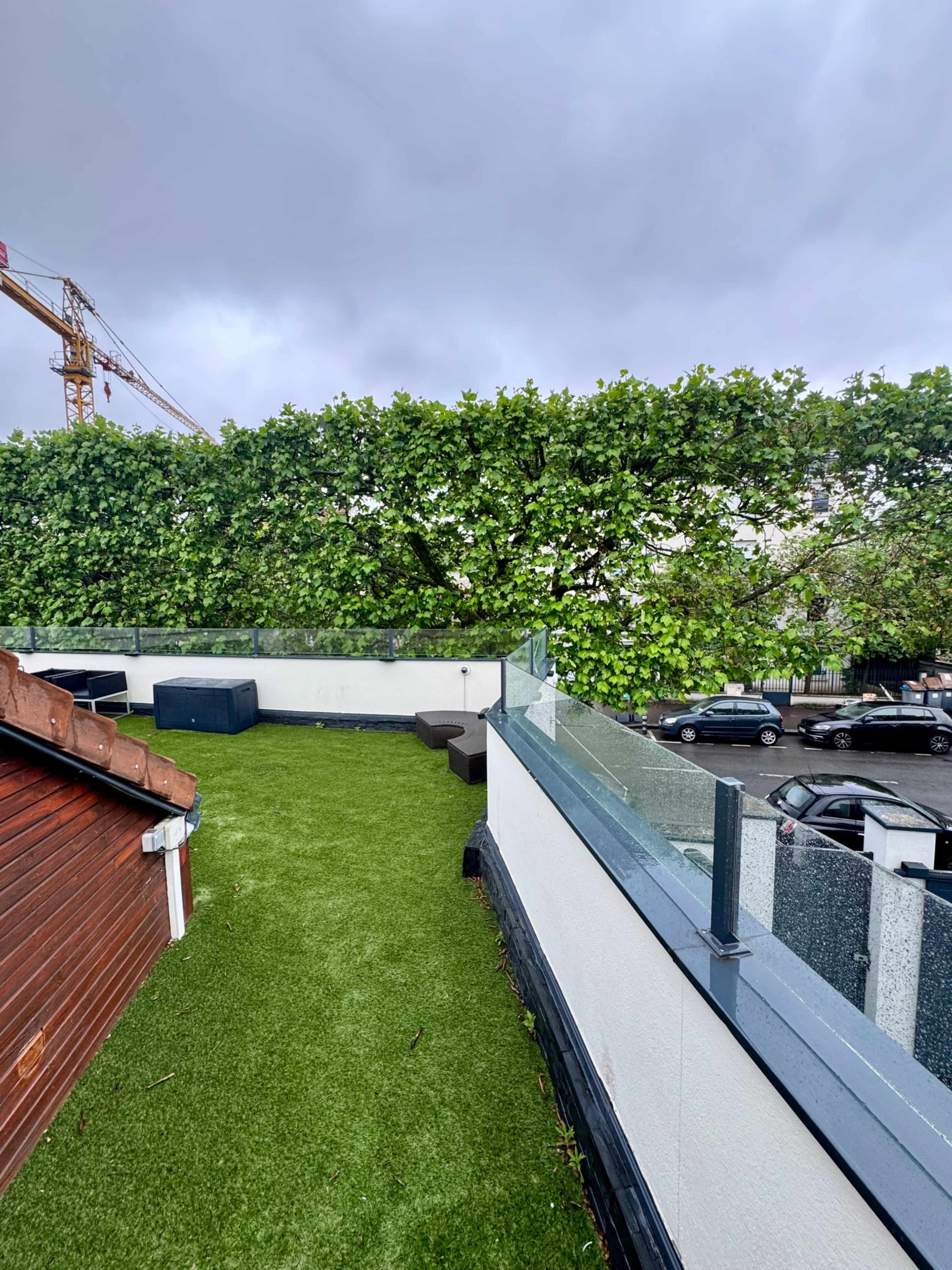 The image shows a rooftop terrace with artificial grass and seating, bordered by a tall hedge, under a cloudy sky.