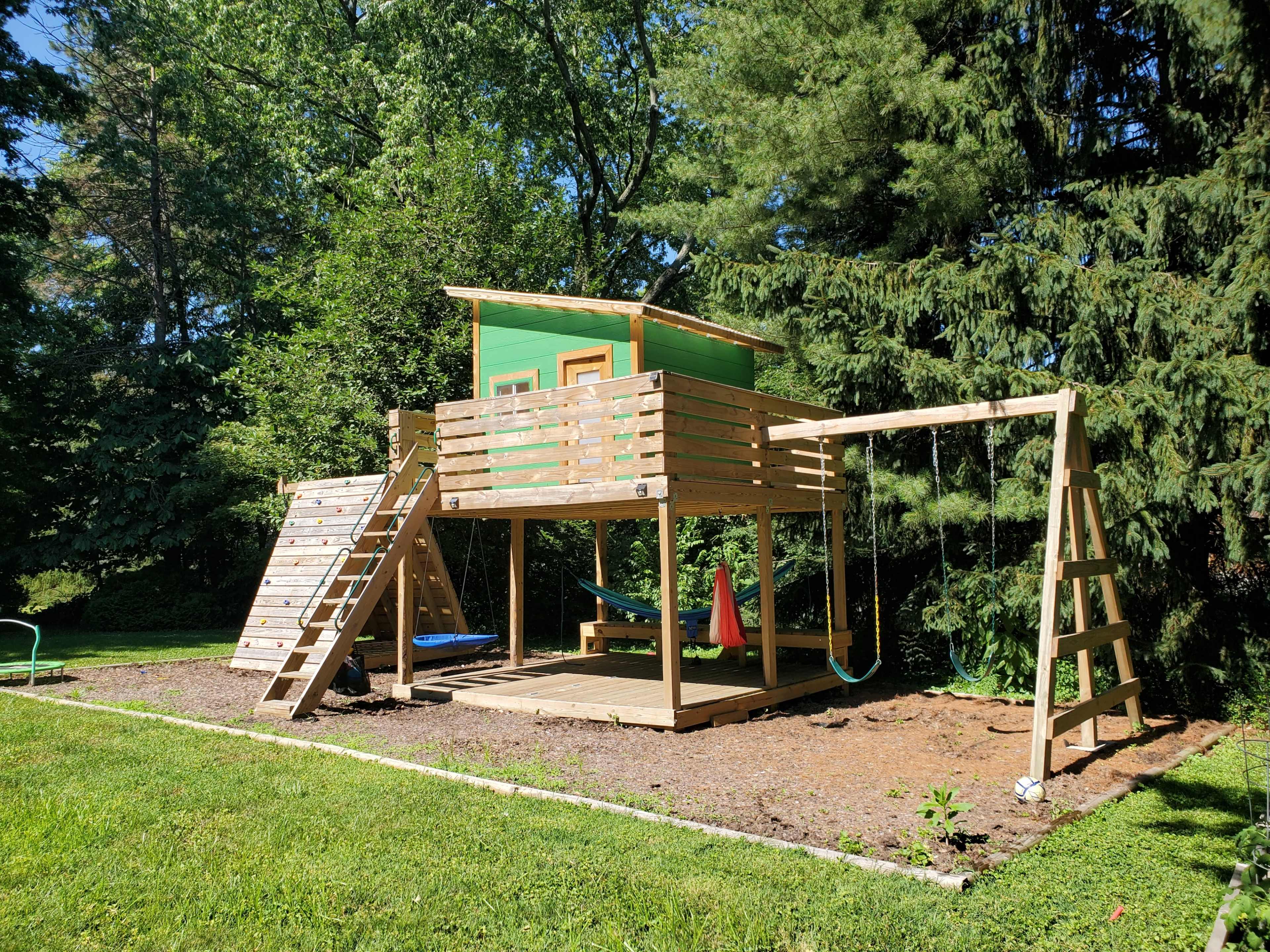 A wooden treehouse with a green roof and a swing set is situated in a grassy area surrounded by trees.