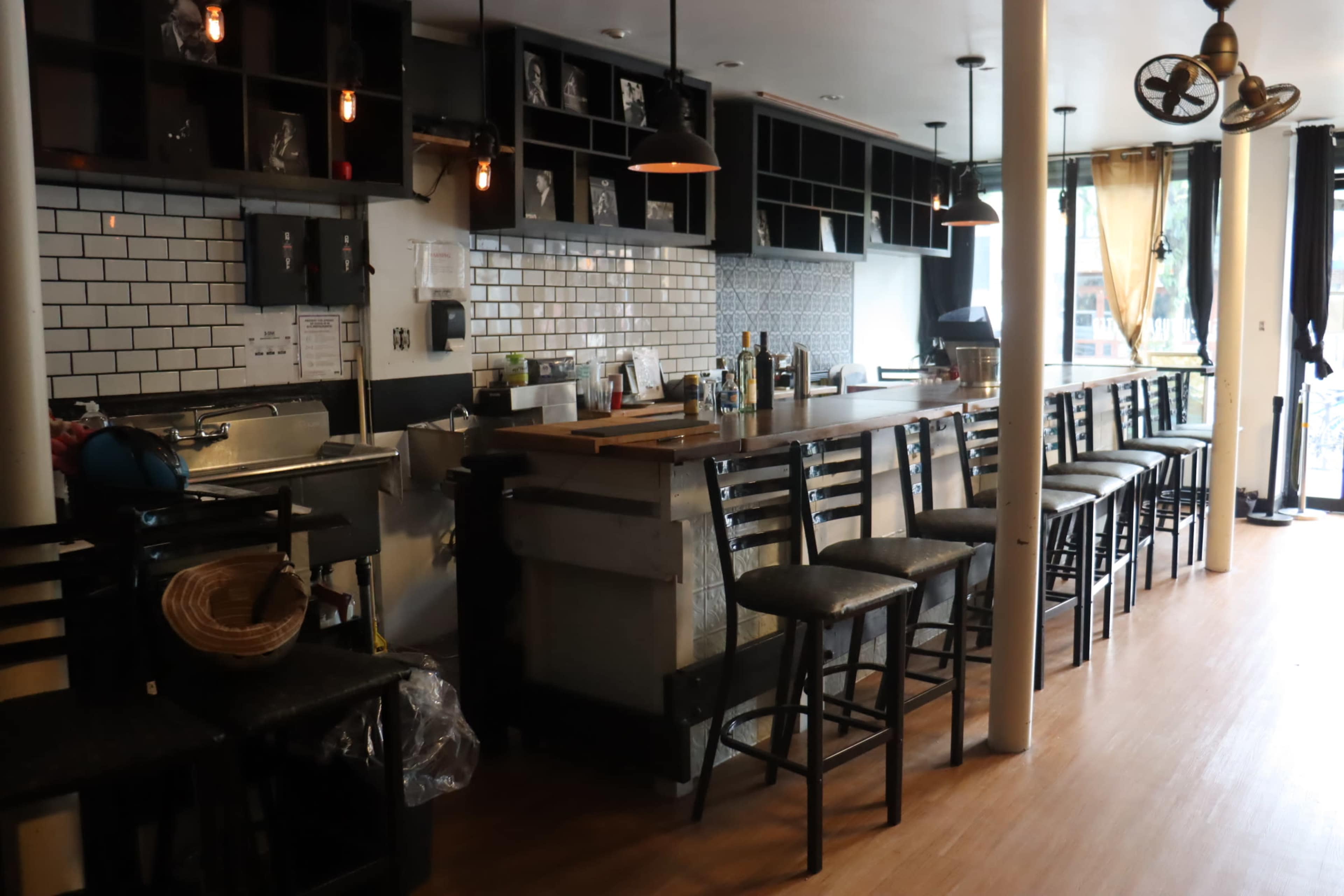 A partially empty bar area with a long countertop, high stools, and a white-tiled wall above the bar shelves.