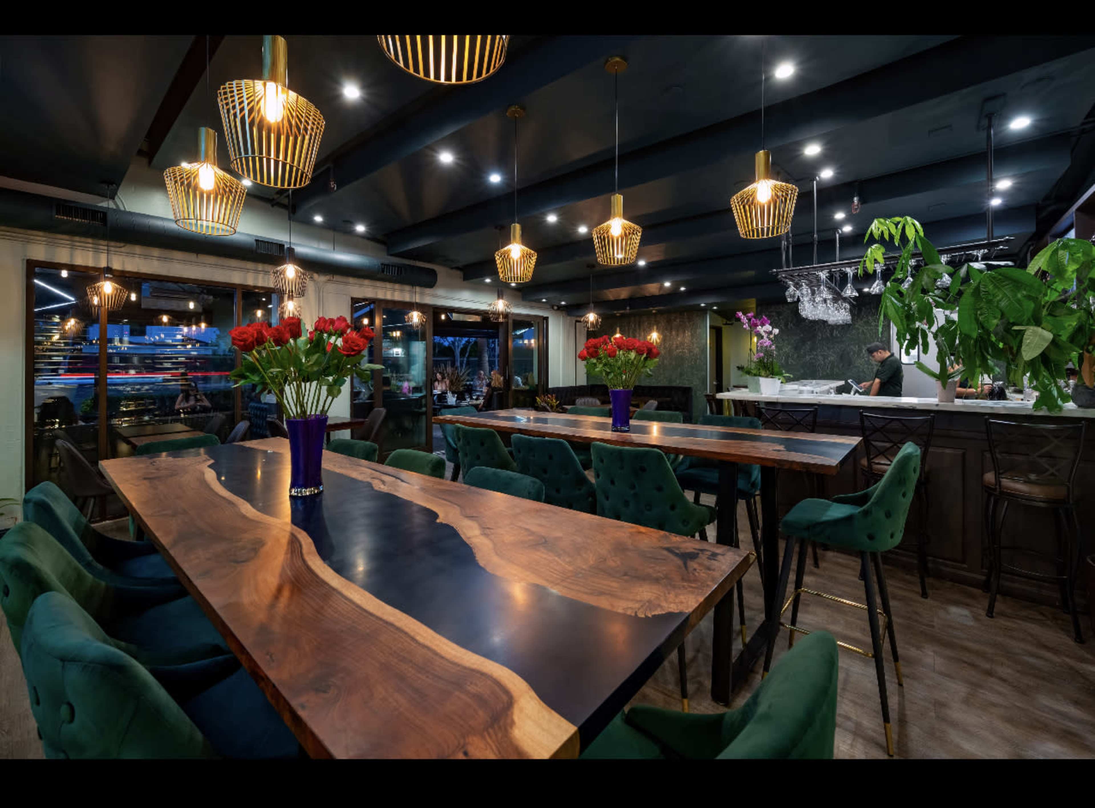 The image shows a modern restaurant interior featuring long wooden tables, green velvet chairs, and pendant lights, with floral arrangements on the tables and a kitchen visible in the background.
