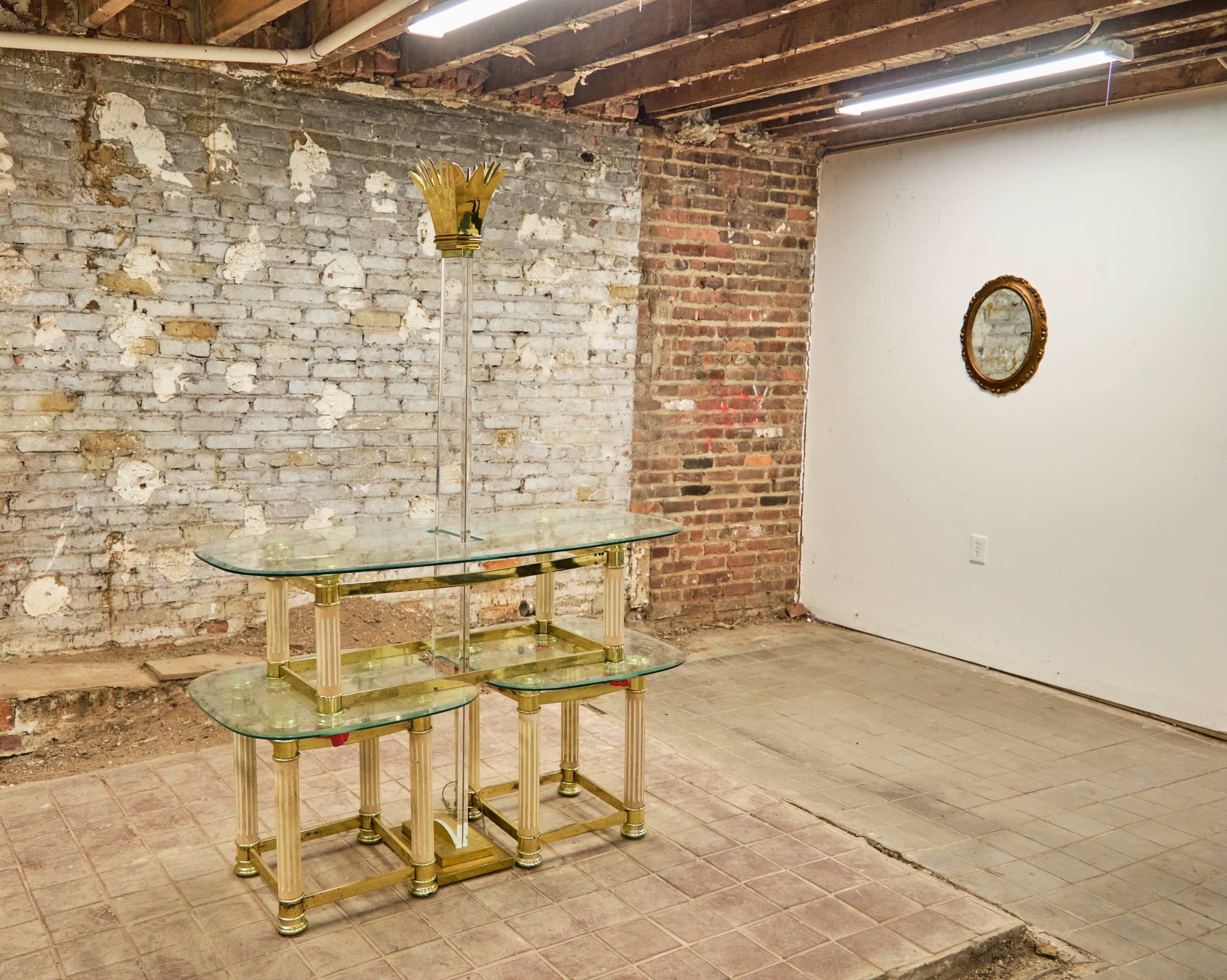 The image shows a glass-top table with a decorative base positioned in an unfinished room with exposed brick walls and a mirror on the opposite wall.