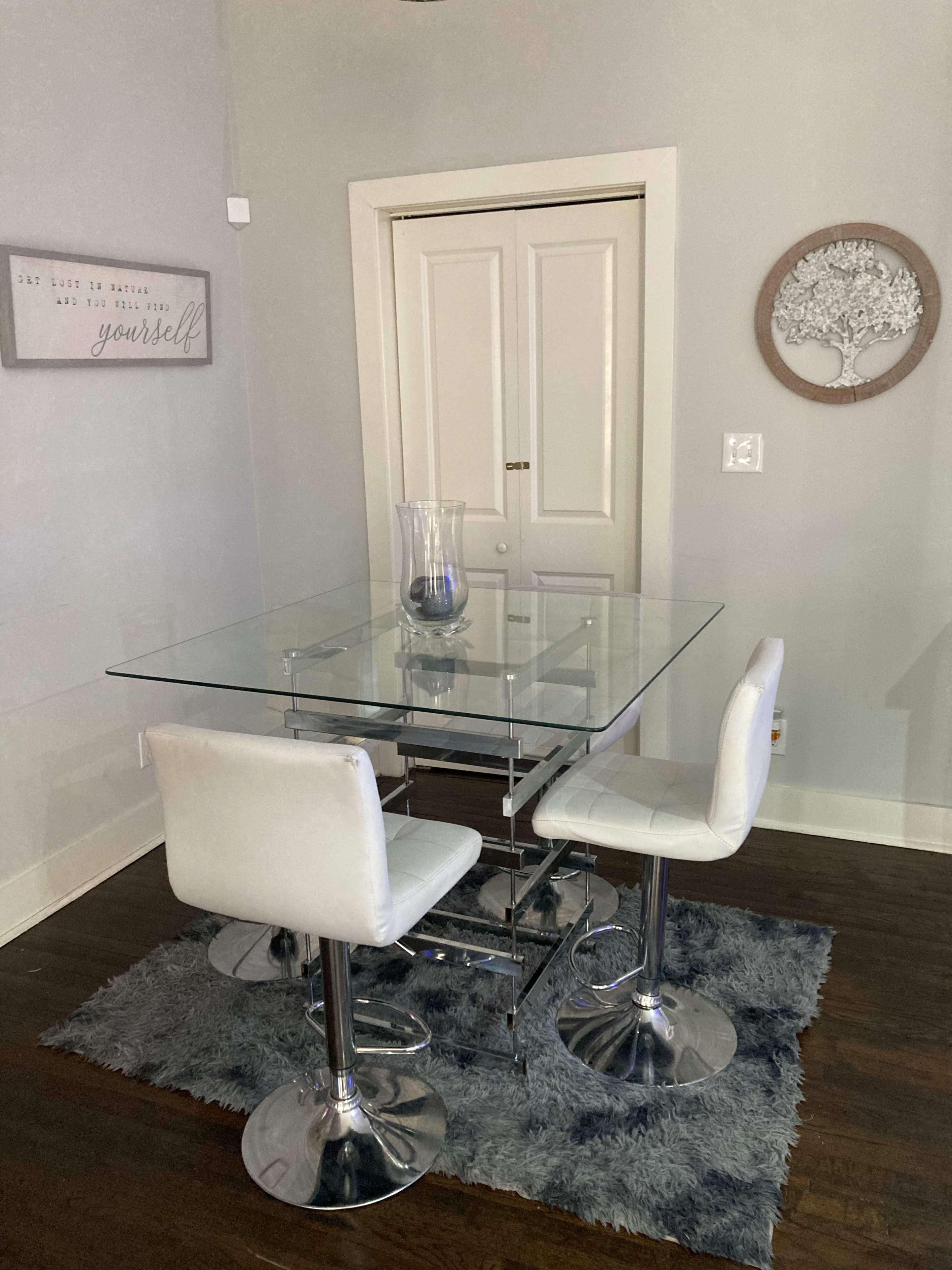 A glass dining table with two white chairs is set on a gray area rug in a room with light gray walls and a closed door in the background.