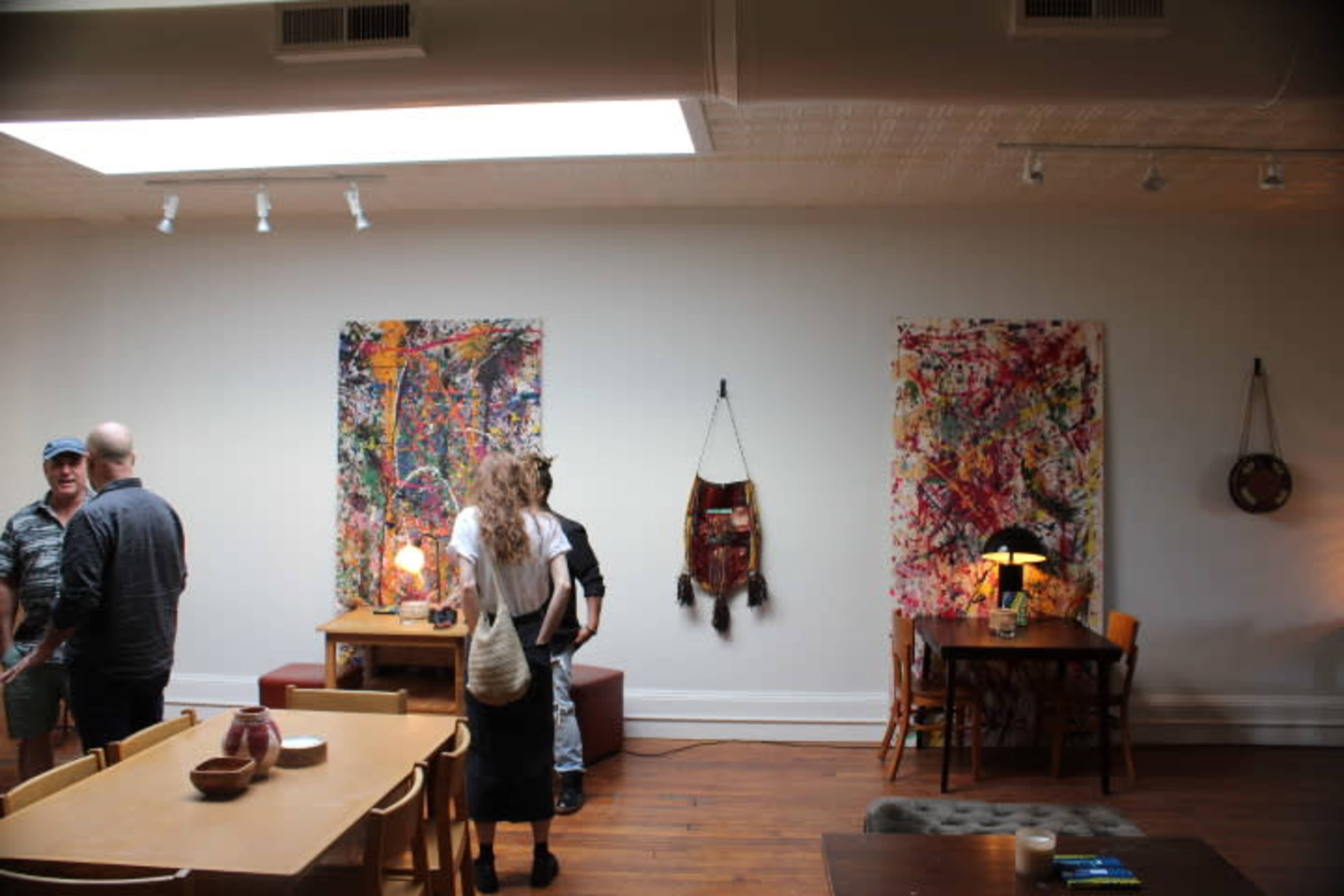 Two groups of people are engaged in conversation in an art gallery adorned with colorful abstract paintings, wooden tables, and soft lighting.