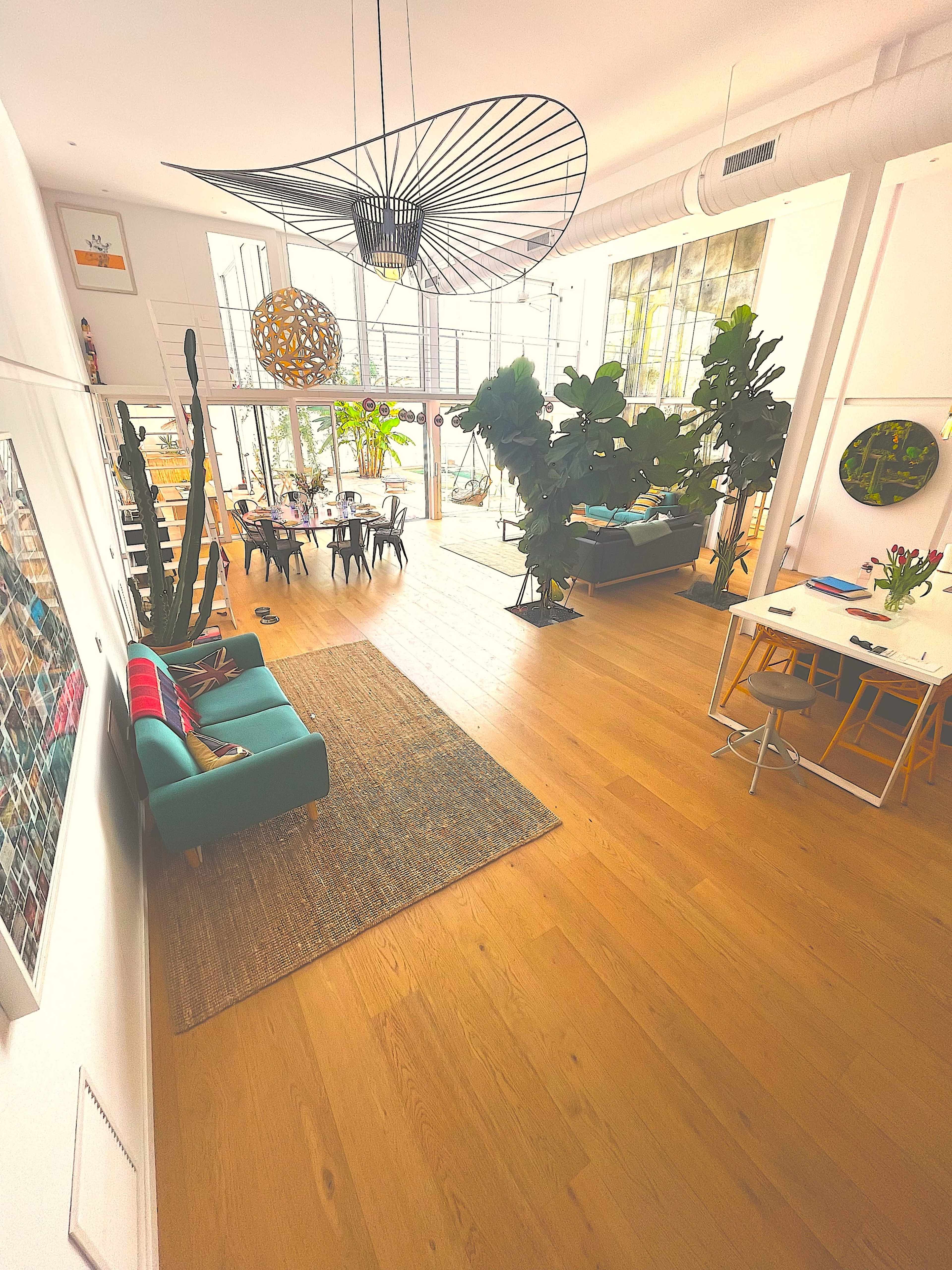 The image shows a bright, modern interior featuring a spacious living area with large windows, various plants, and contemporary furniture.