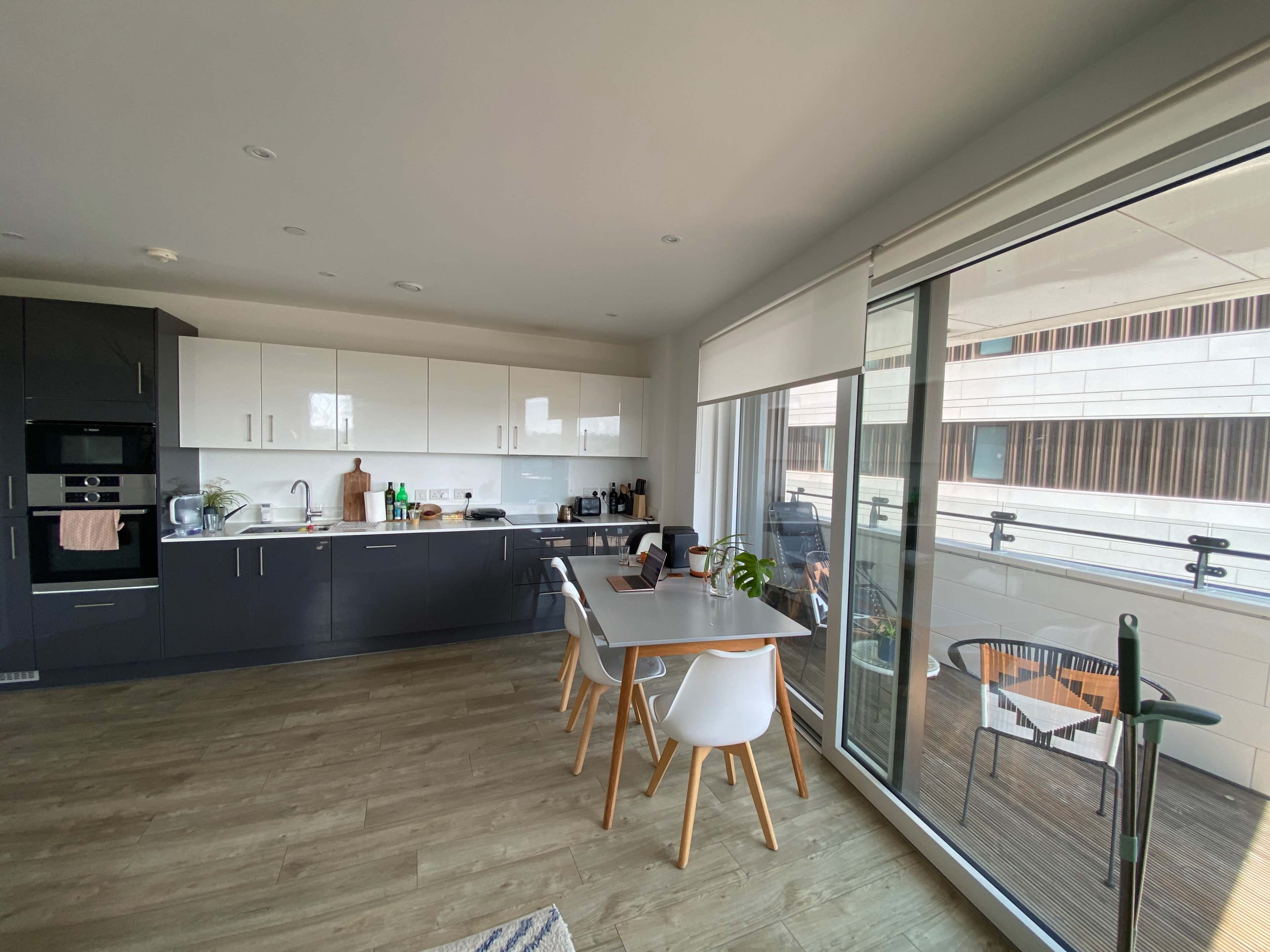 The image shows a modern kitchen with dark cabinets, a white countertop, and a dining table set against a backdrop of a balcony.