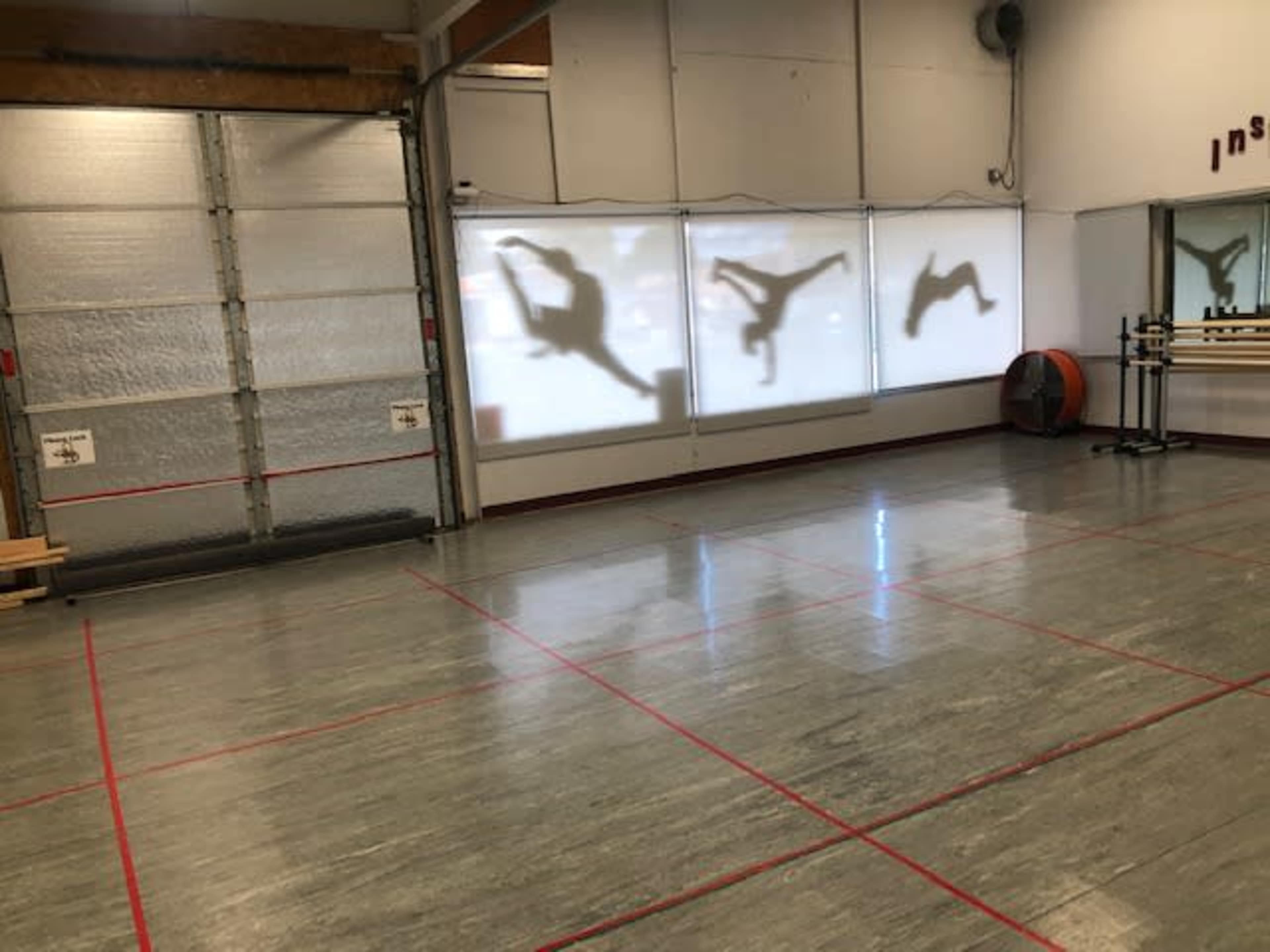 An empty dance studio with a smooth wooden floor and shadows of dancers projected onto a wall.