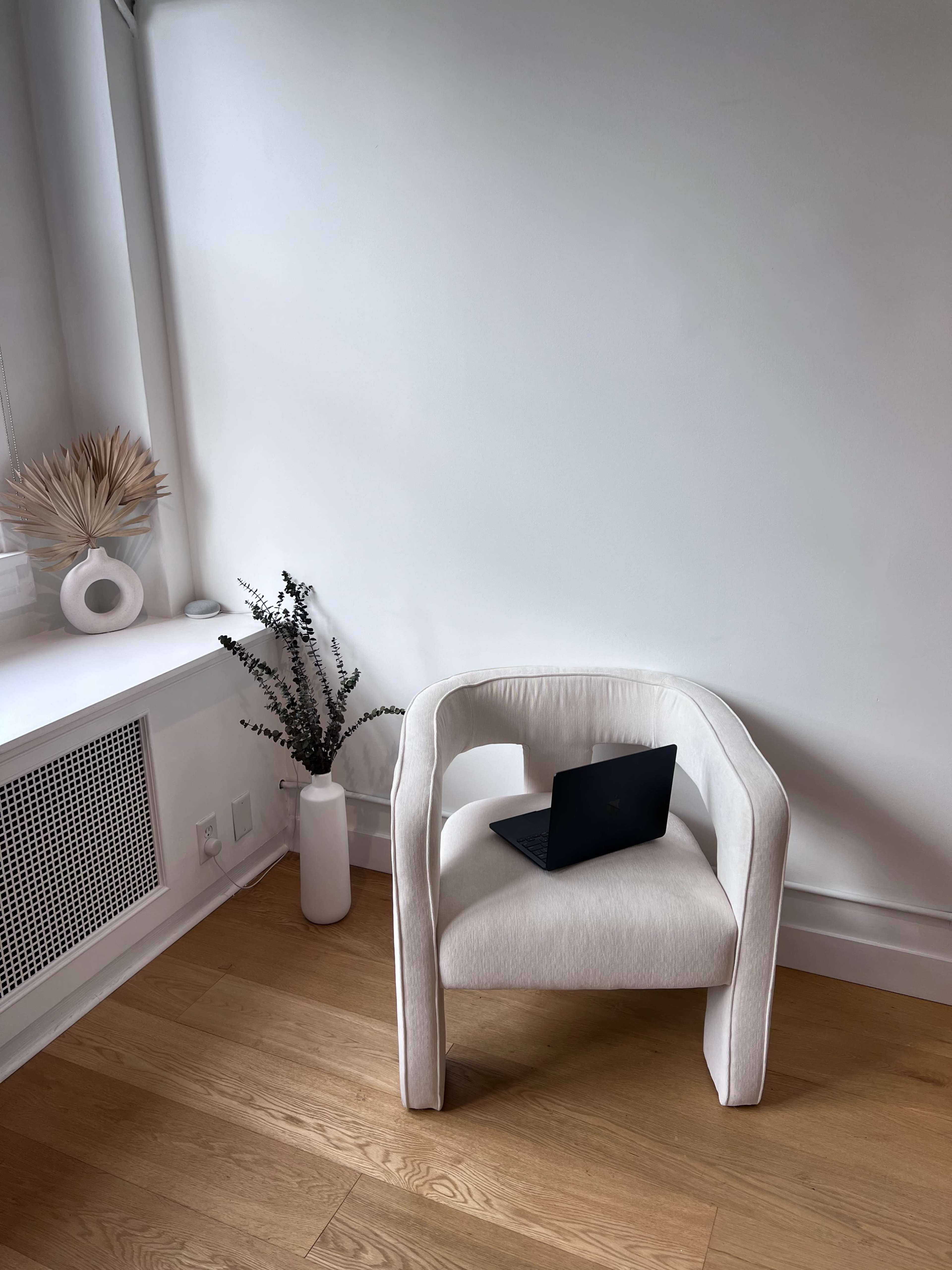 A light-colored chair with a laptop on it sits beside a decorative plant in a minimalist room with wooden flooring.