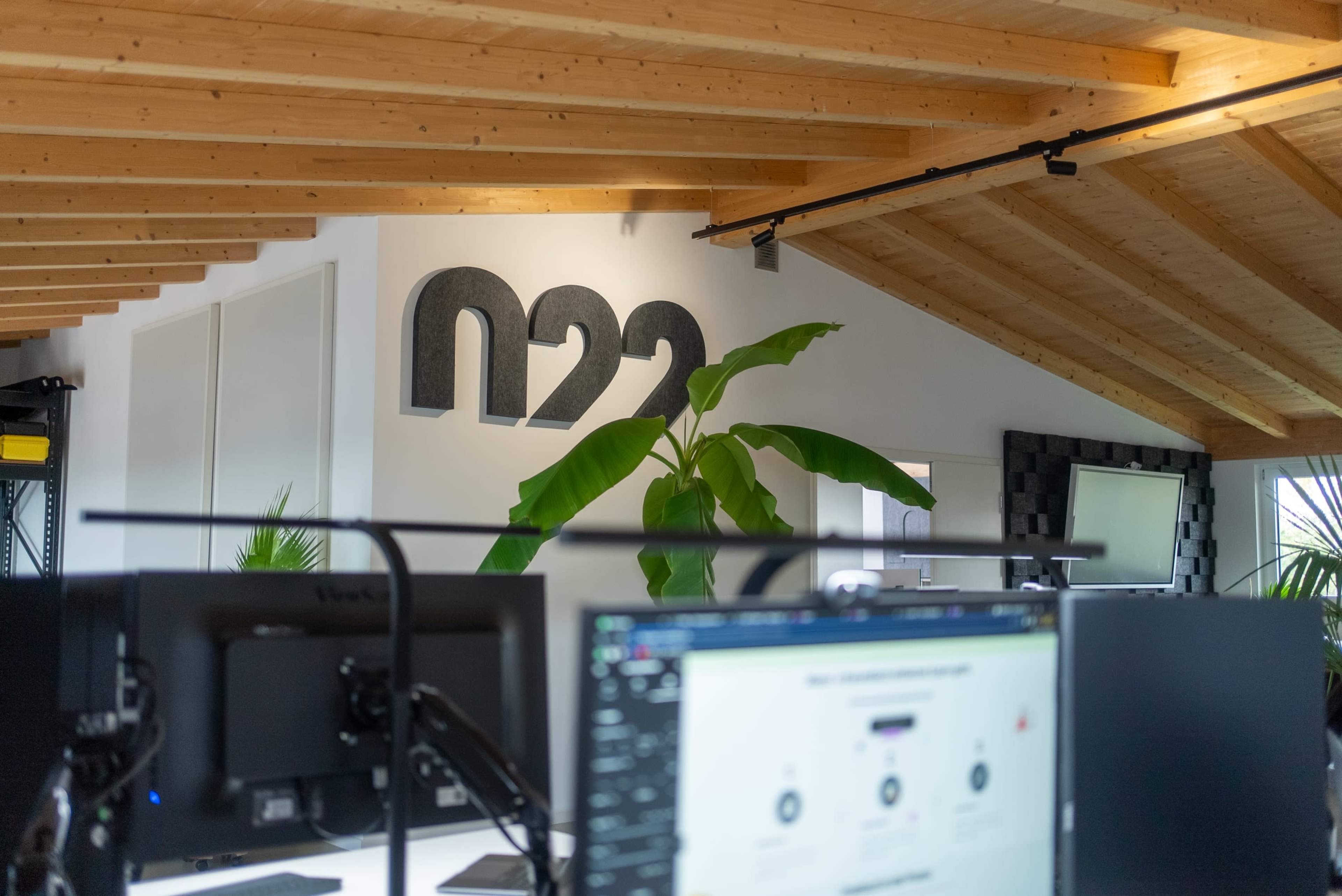 The image shows an office space with a wooden ceiling, featuring a large plant and the letters "n22" mounted on the wall.