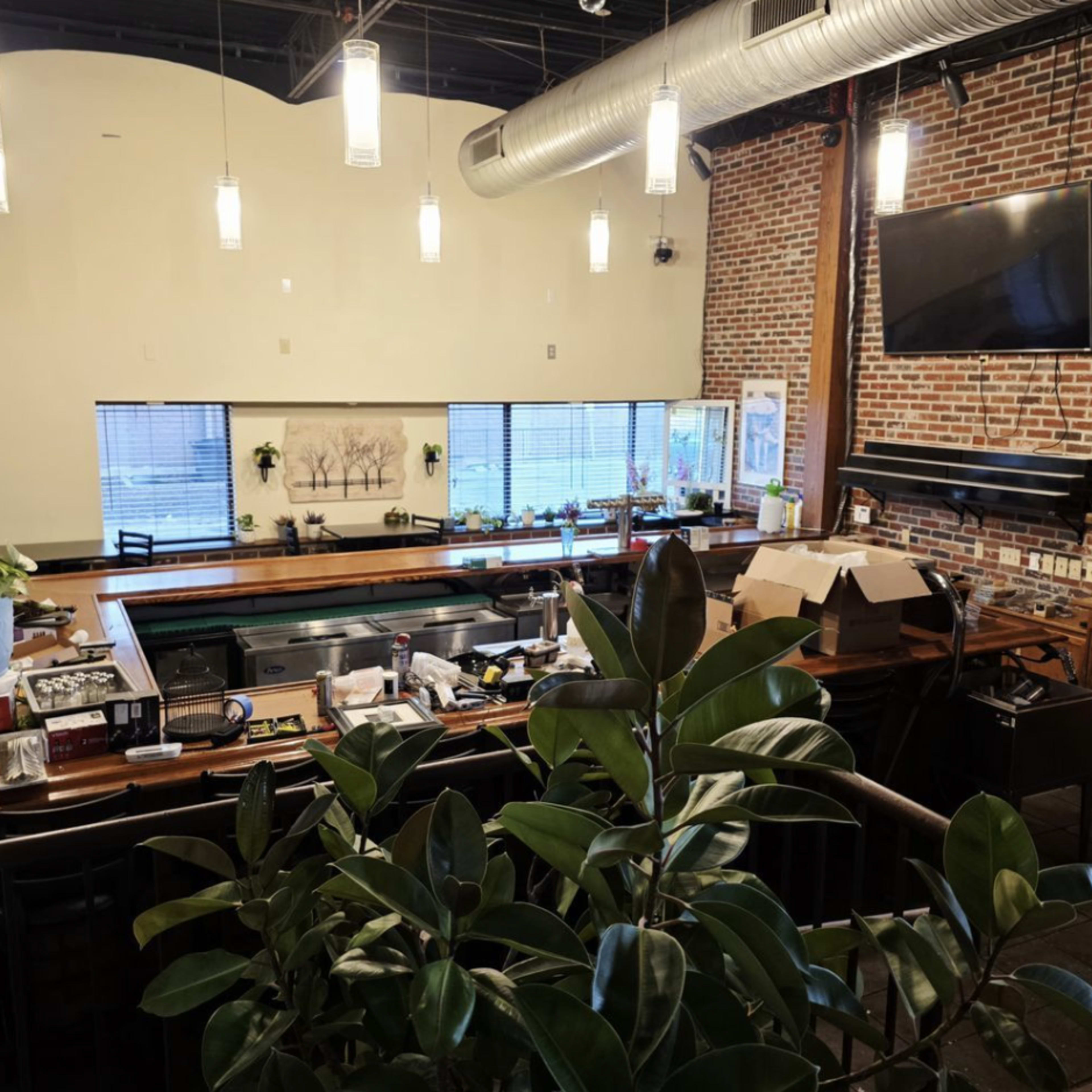 The image shows a bar area with a wooden countertop and a large window featuring planters, overlooking a clean and modern space with exposed brick walls and overhead lighting.