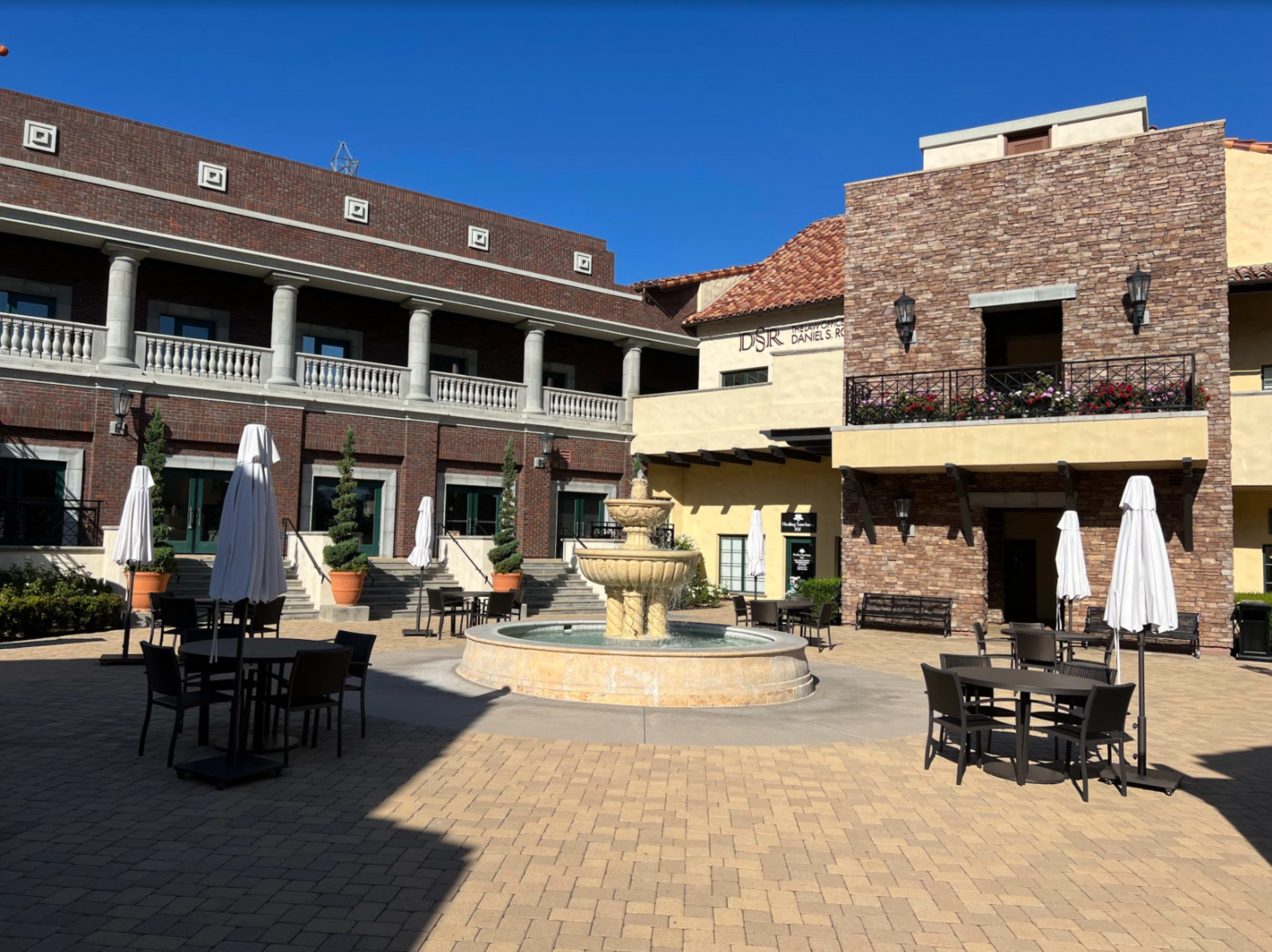 A courtyard featuring a central fountain, surrounded by tables and chairs under white umbrellas, with brick and stucco buildings in the background.