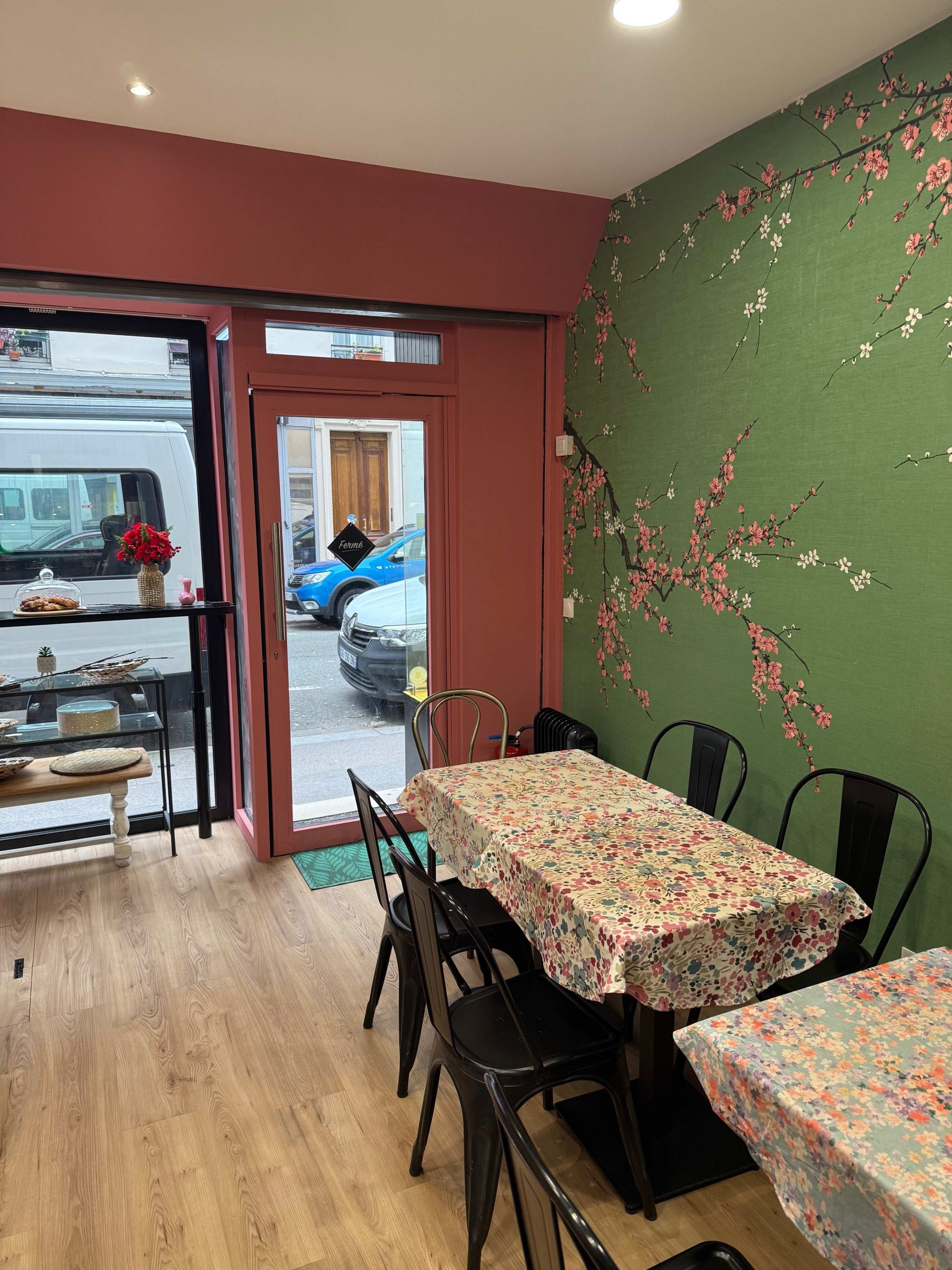 The image shows an indoor café or restaurant with floral-patterned tables, black chairs, and a wall adorned with a green floral mural, while a view of the street is visible through a large window.