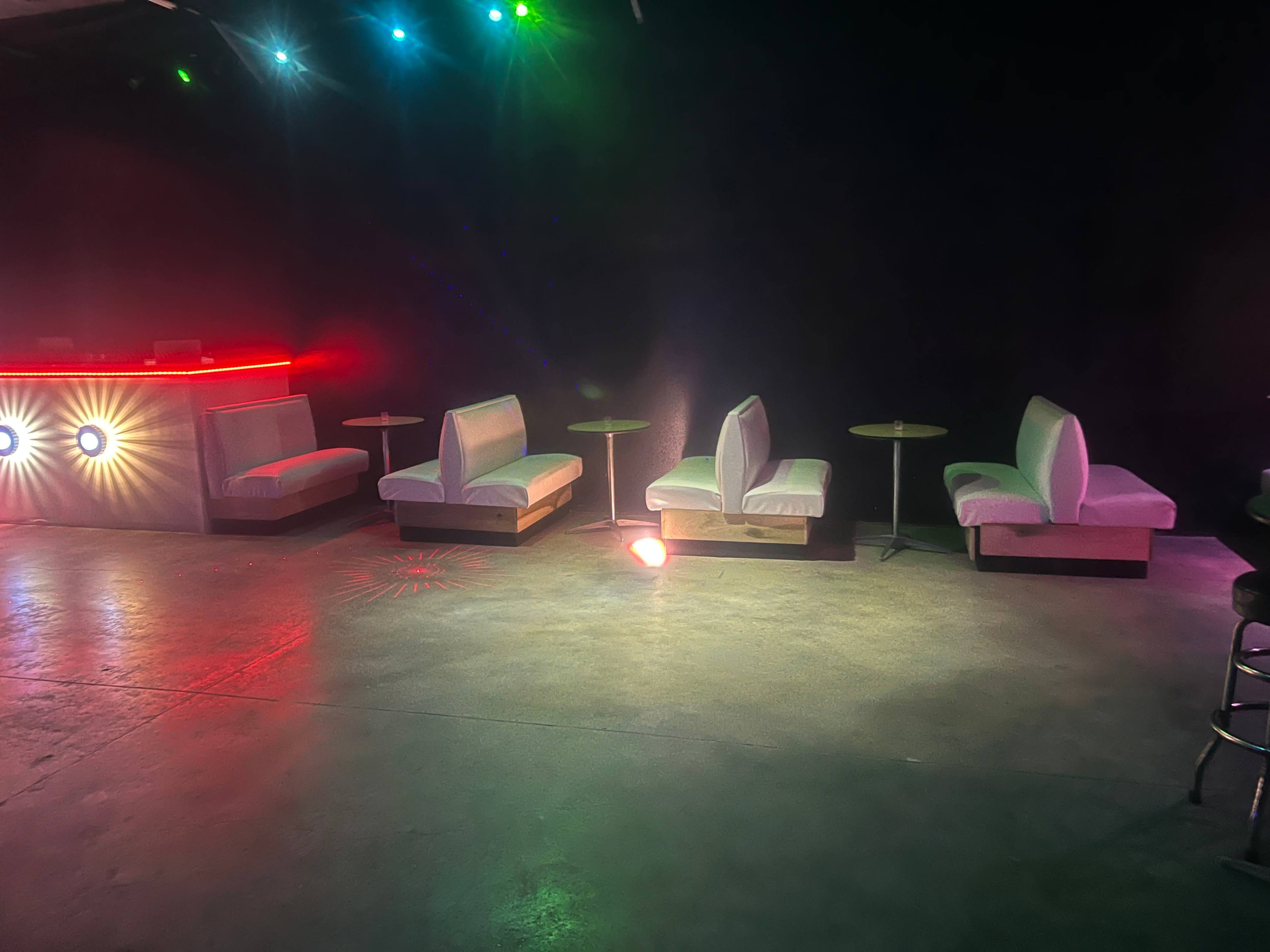 The image shows a dimly lit lounge area with white sofas and small round tables, illuminated by colorful lights.