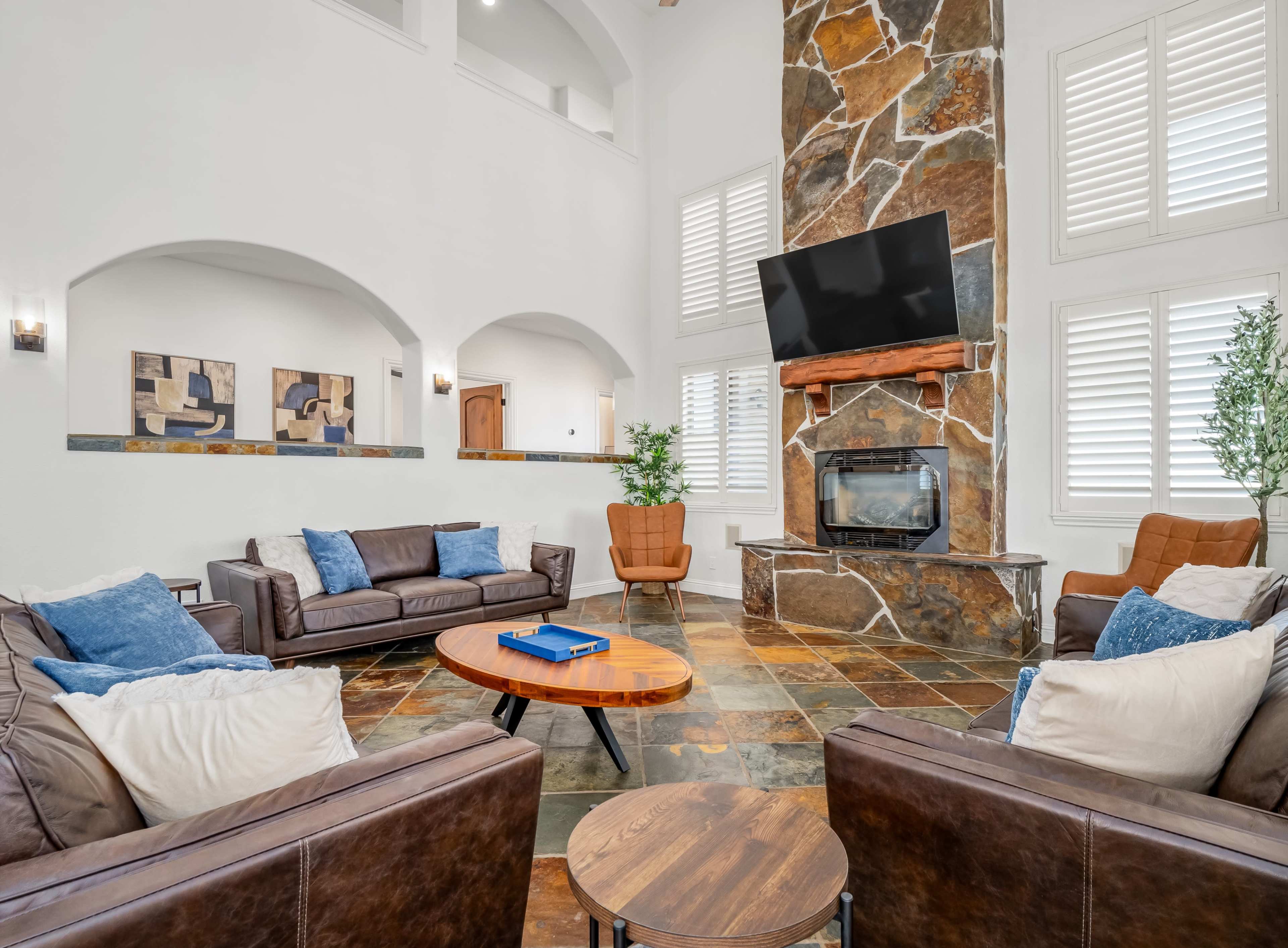 A spacious living room with a stone fireplace, large windows, and leather seating arranged around a circular wooden coffee table.