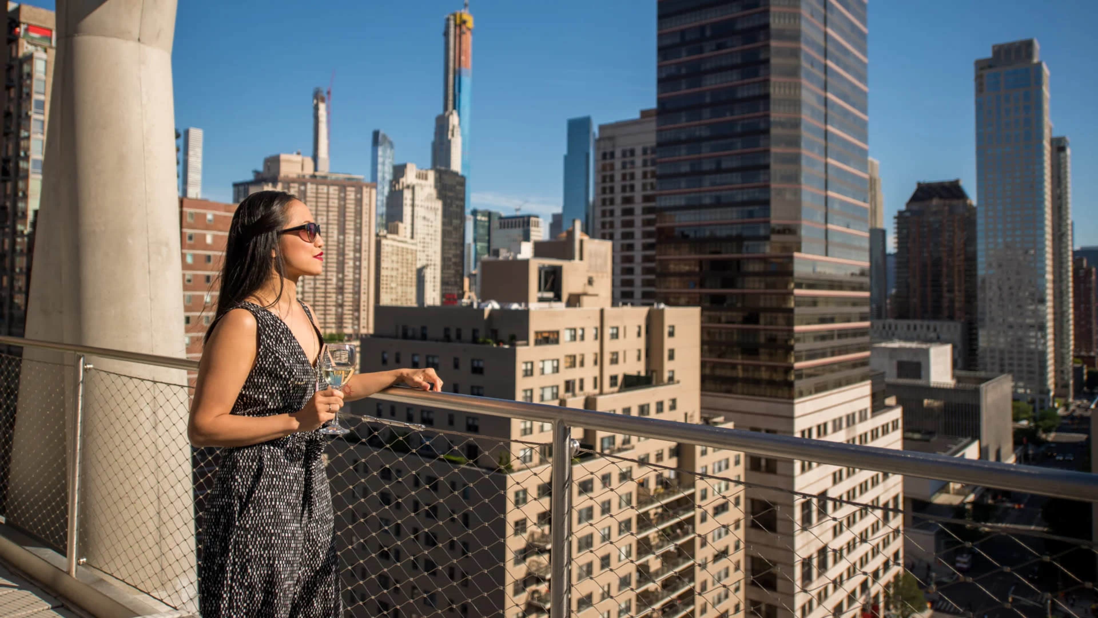 A woman holding a glass stands on a balcony overlooking a city skyline filled with tall buildings and clear blue skies.