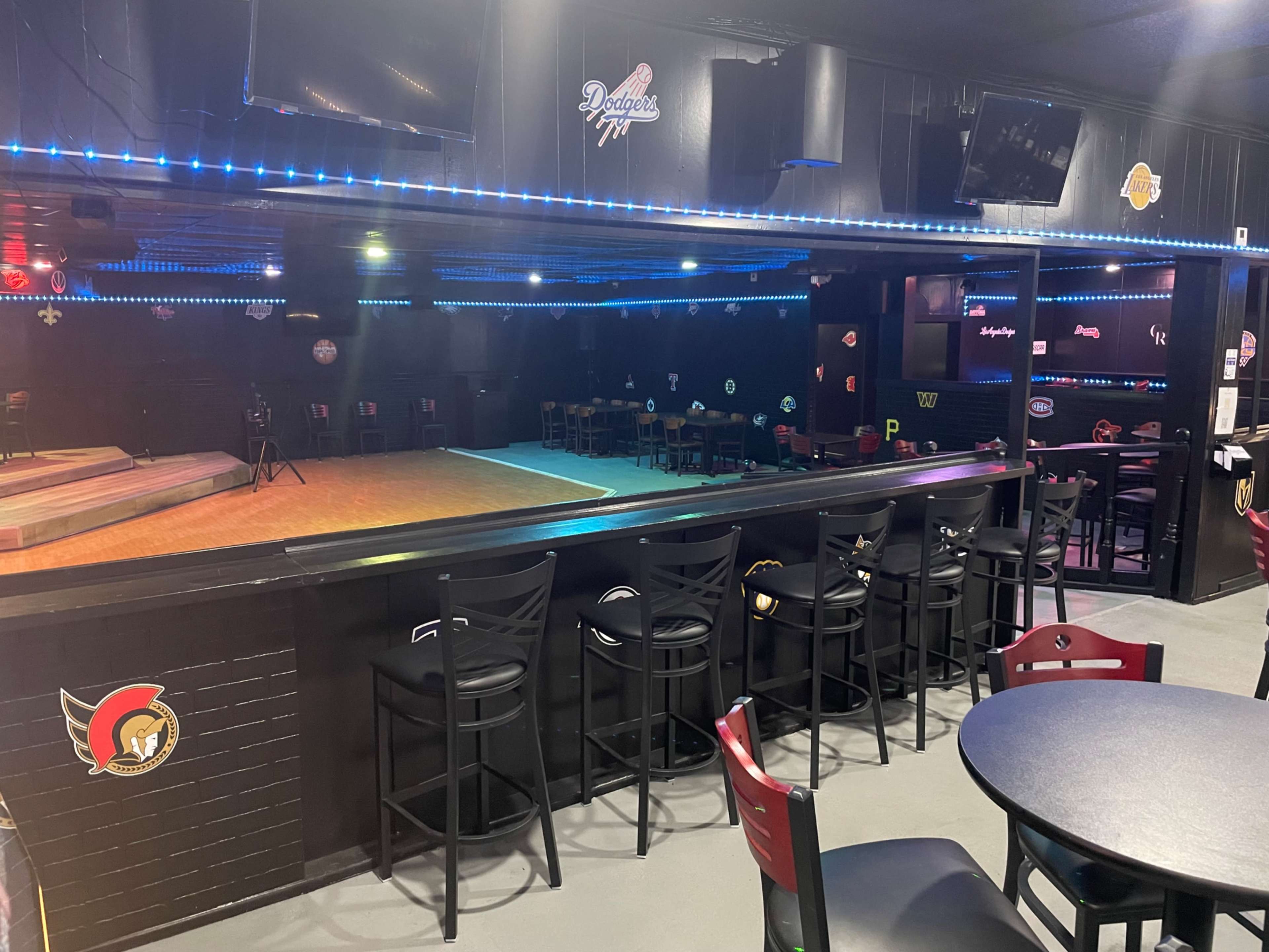 A dimly lit sports bar features a stage area with seating, a bar with stools, and various team logos displayed on the walls.