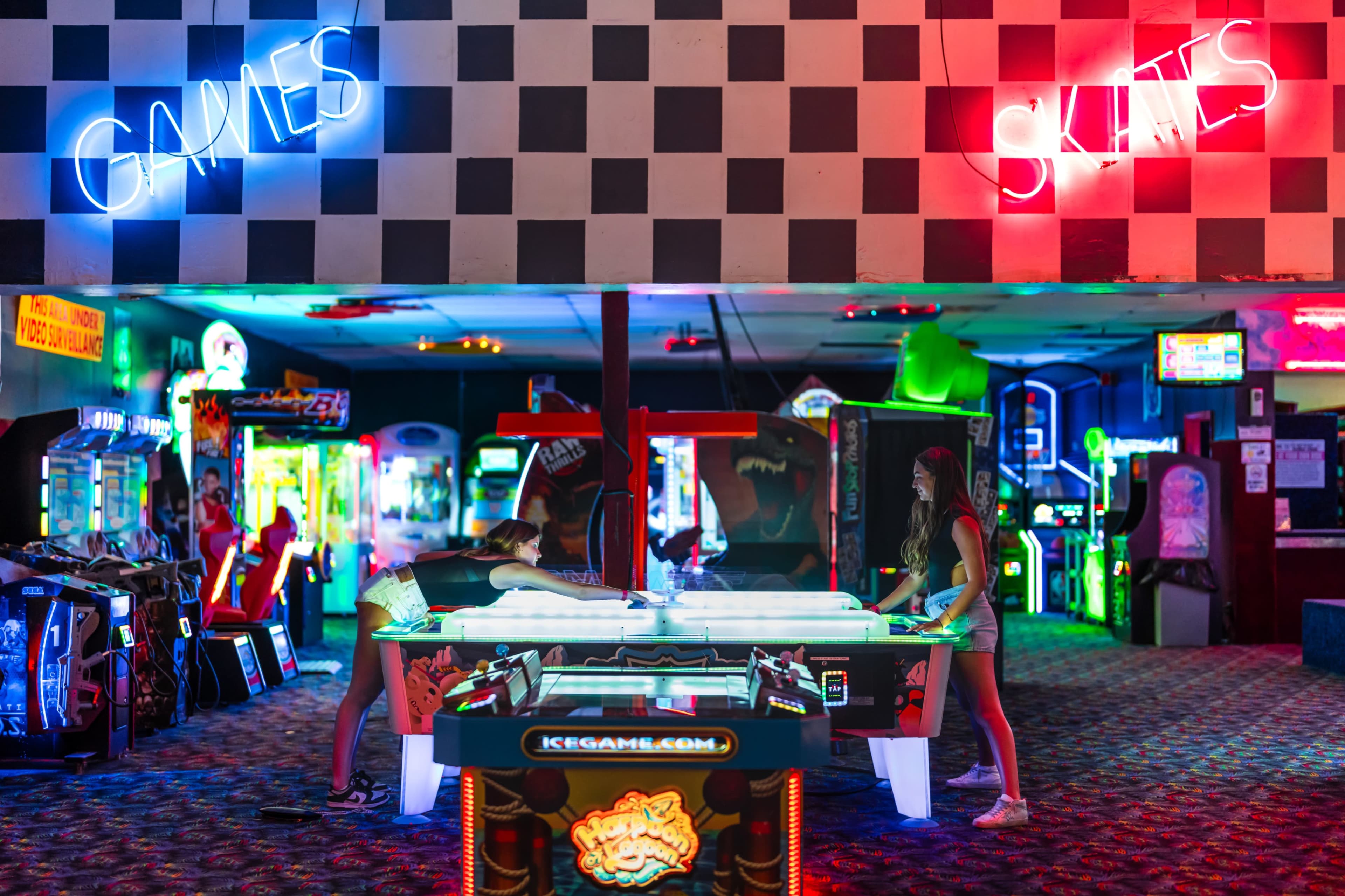 Two women play air hockey in a vibrant arcade filled with various games and neon lights.
