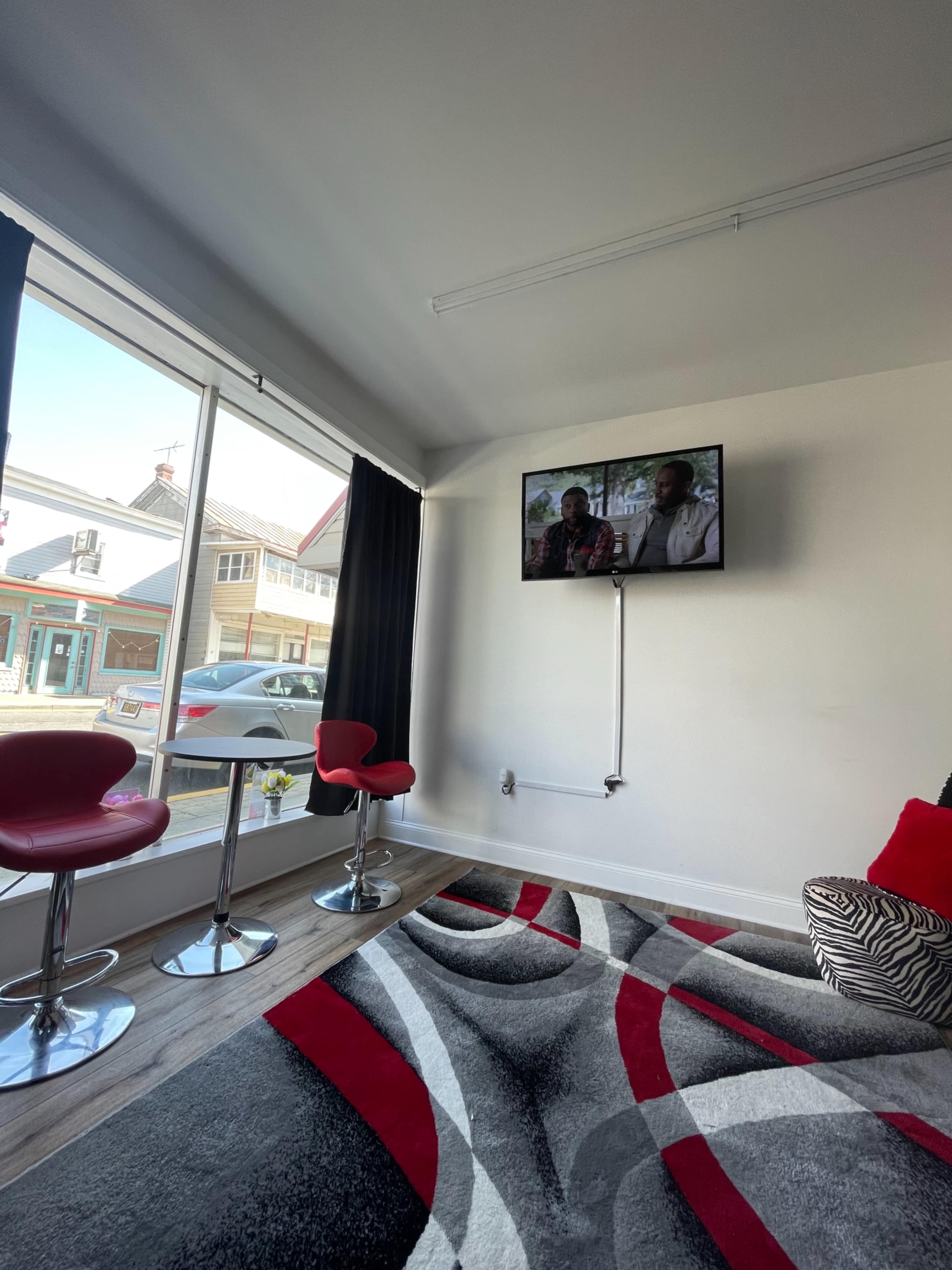 A modern indoor space with two red bar stools in front of a television mounted on the wall, beside a large window and a patterned rug on the floor.