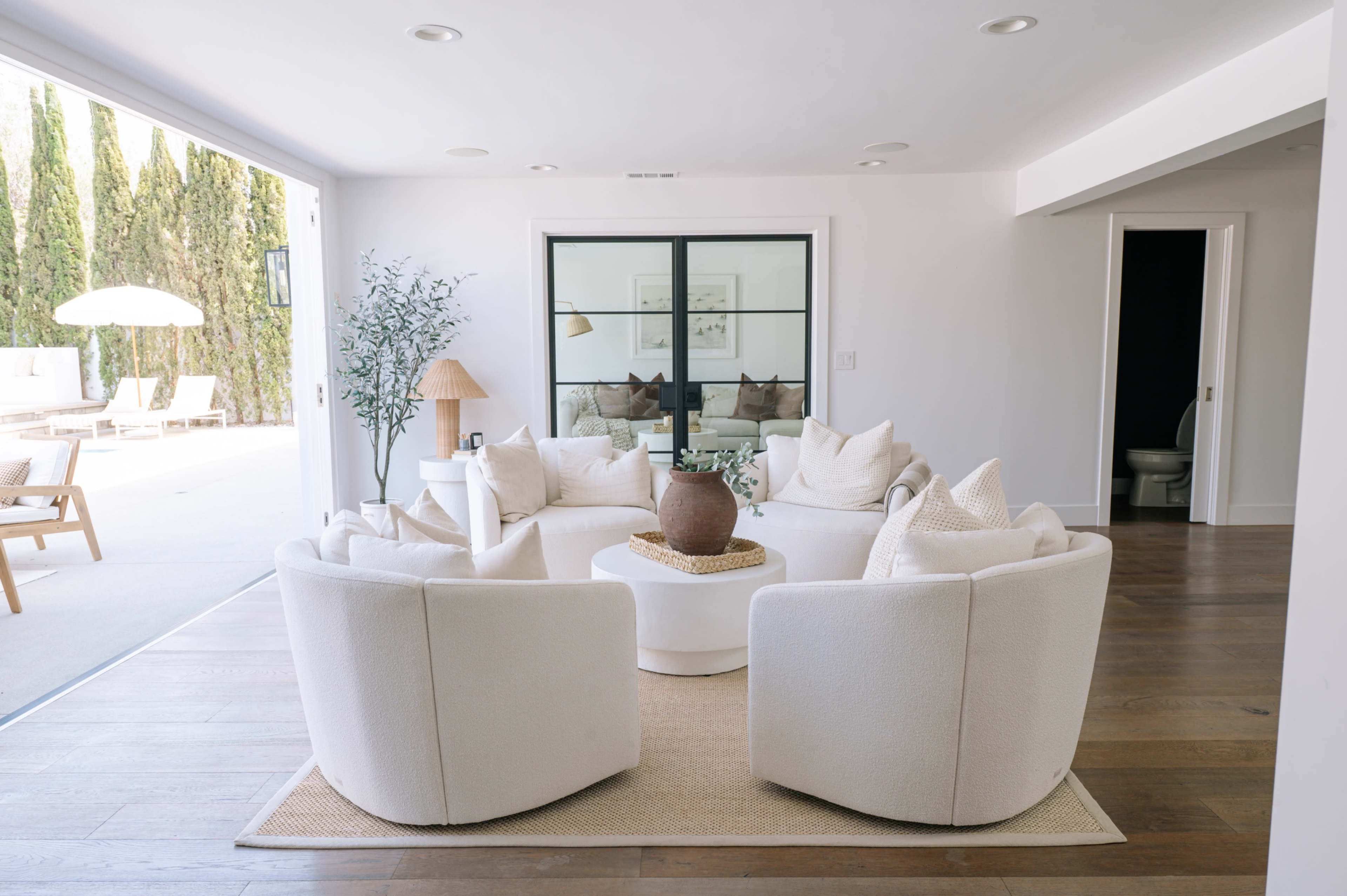 The image shows a modern living room with a circular arrangement of white upholstered sofas around a round coffee table, featuring large windows that overlook a patio with greenery outside.