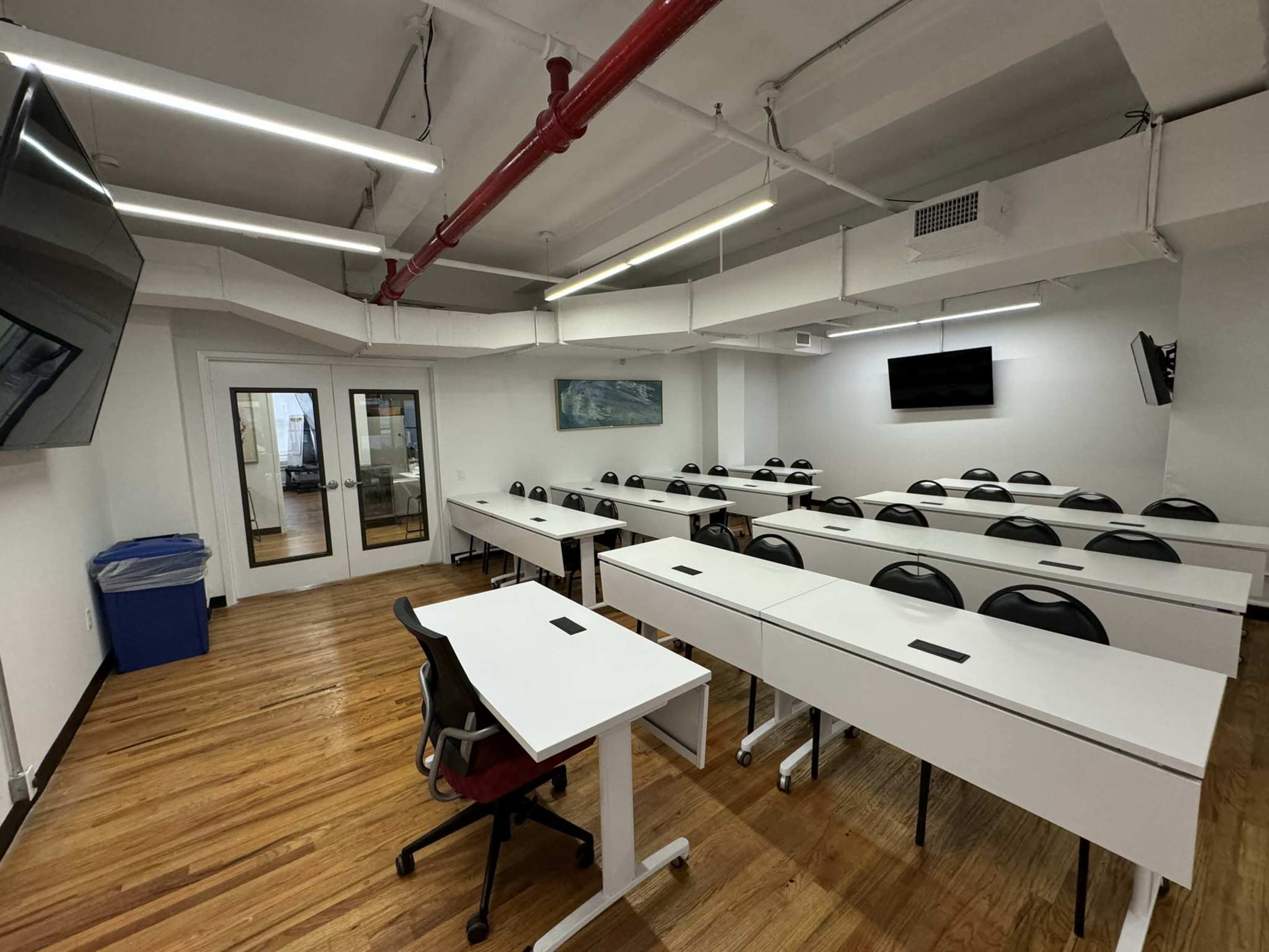 A bright classroom features multiple rows of desks and chairs, with two large screens on the walls and windows leading to an adjacent space.