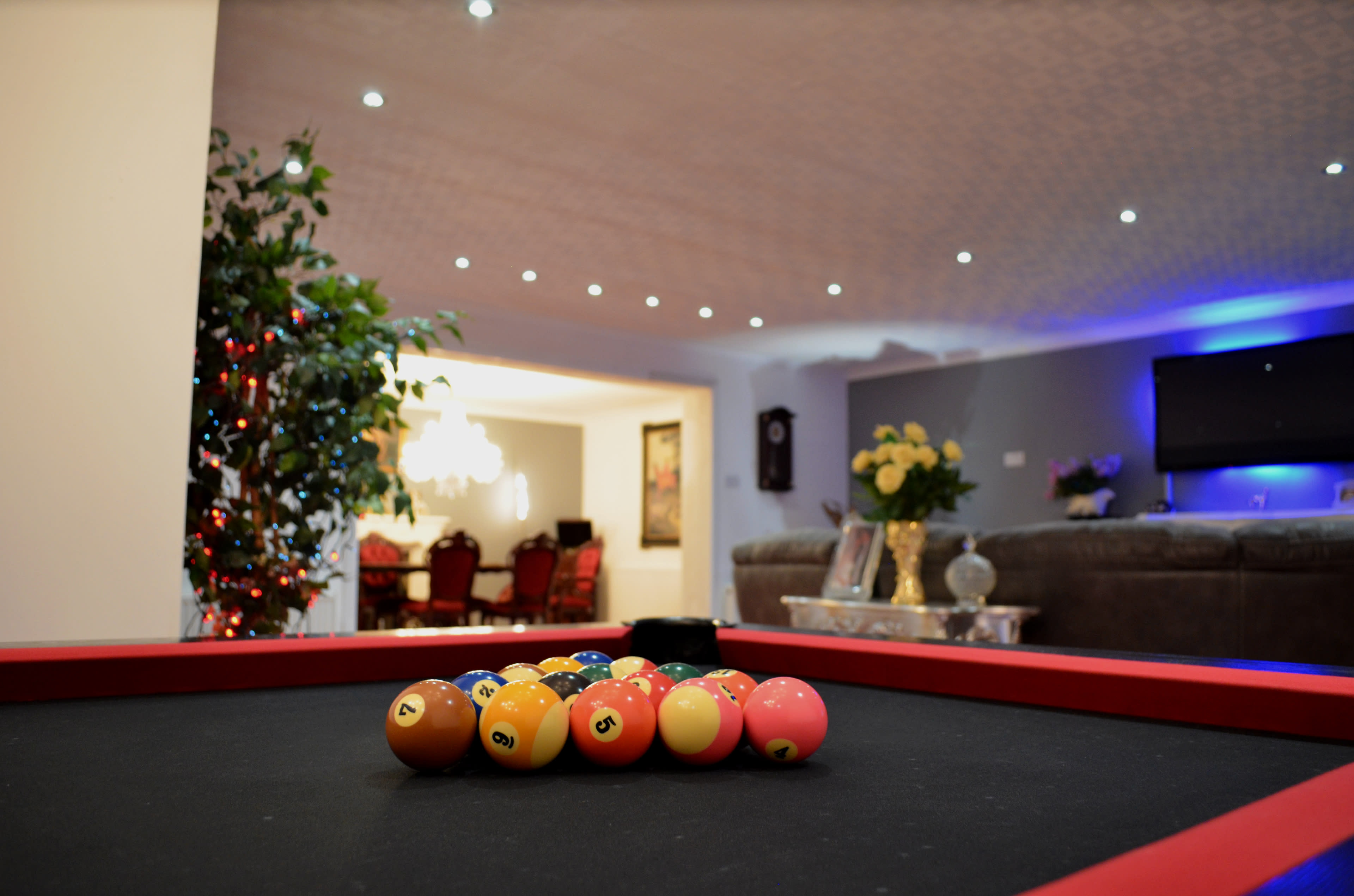 A billiard table with balls arranged in a triangle is set in a brightly lit room that features potted plants and a view of a living area with a couch and dining table.