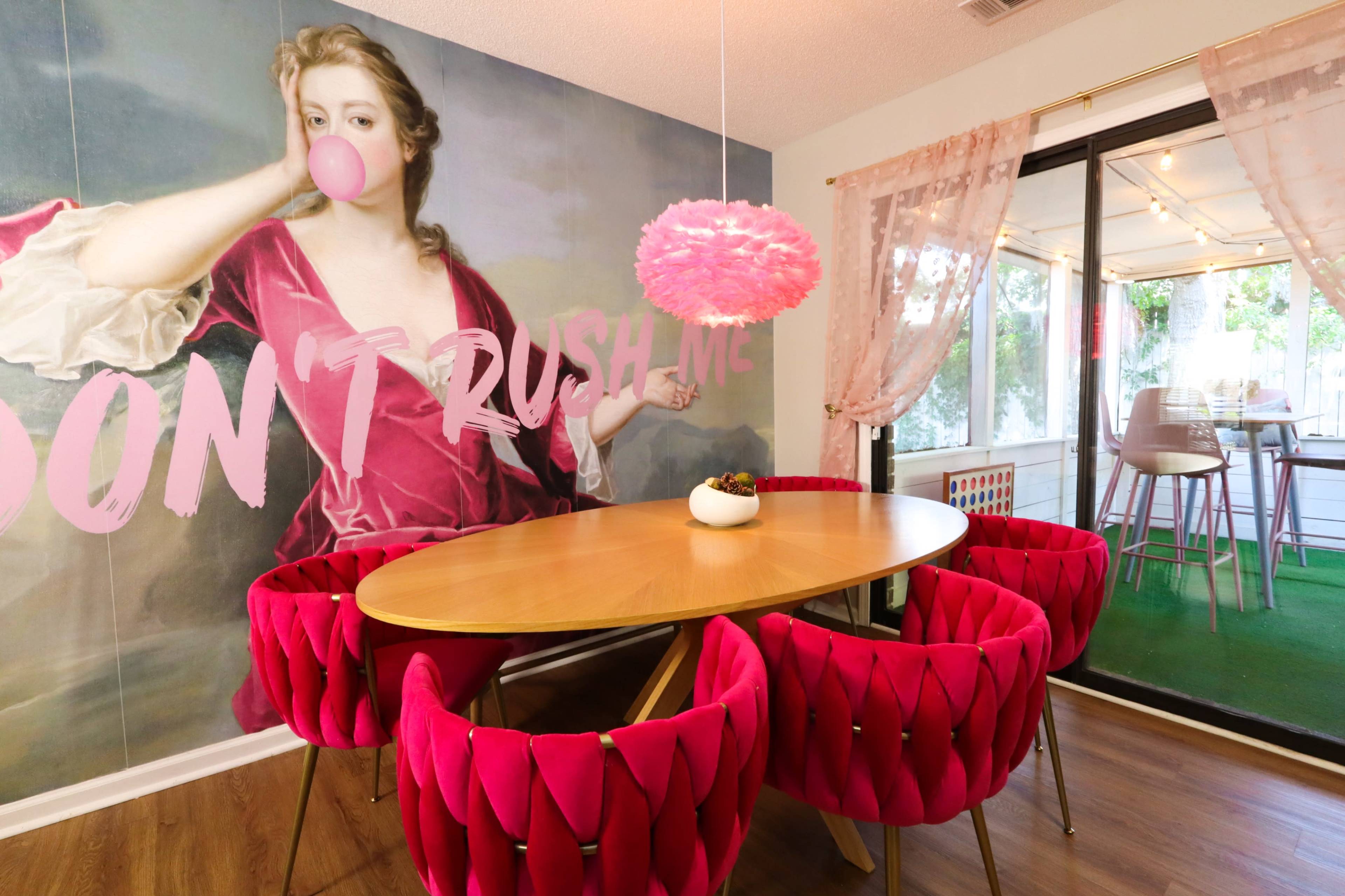 A dining area features a round wooden table surrounded by pink tufted chairs, with a large mural of a woman blowing a bubble gum and the words "DON'T RUSH ME" on the wall.