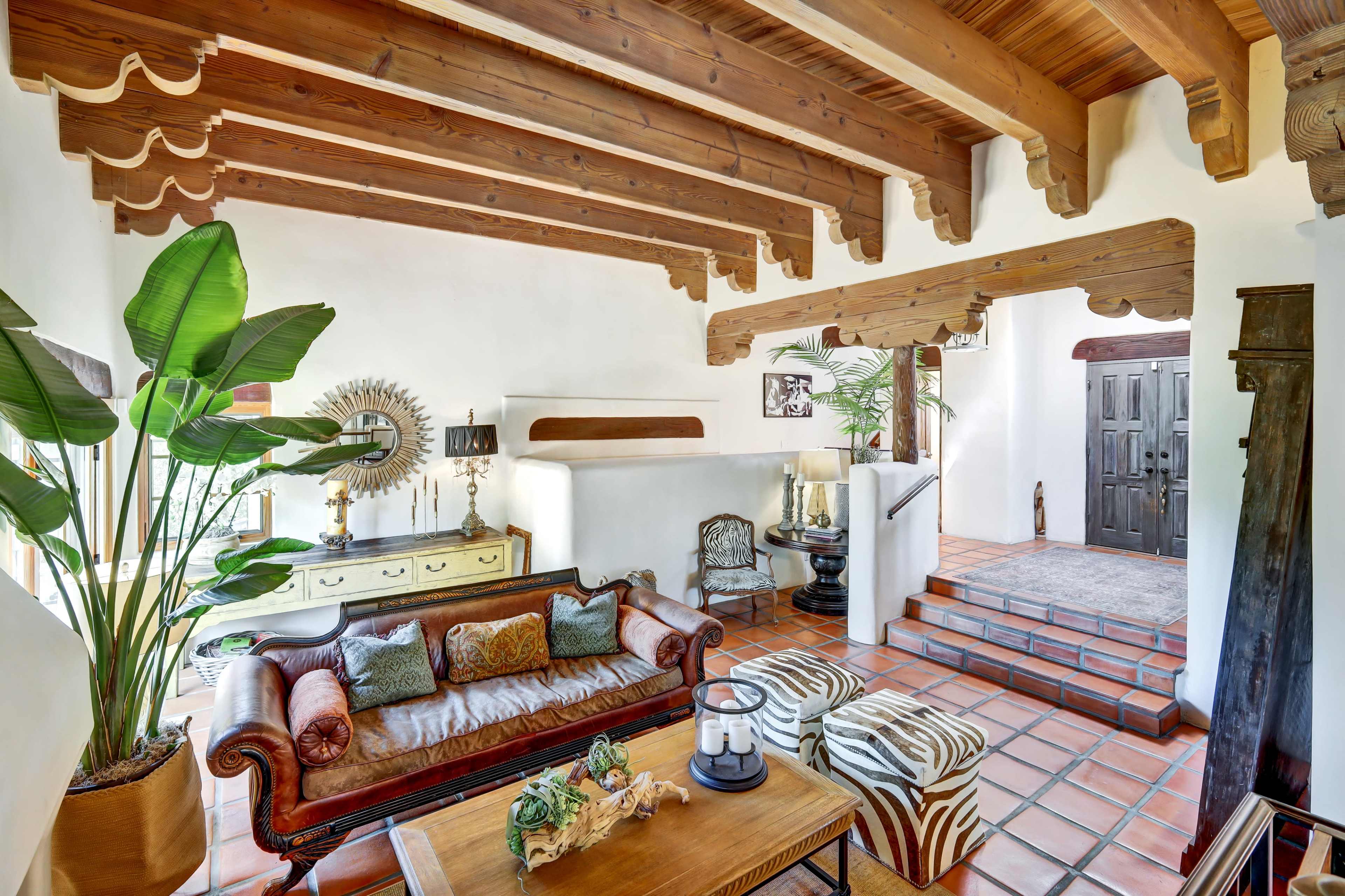 The image shows a living room featuring wooden beams, a leather sofa, decorative plants, and an entrance leading to a staircase.