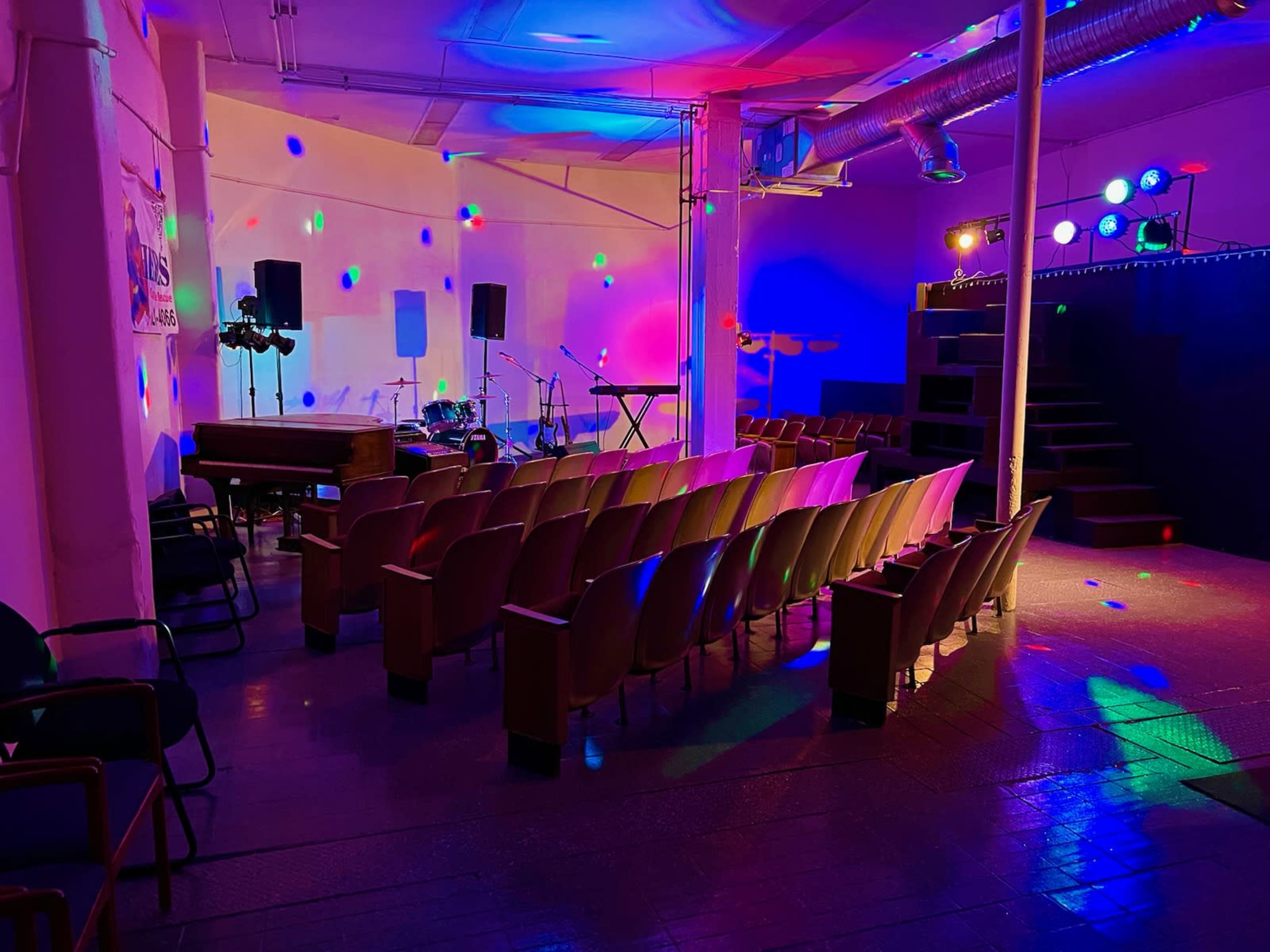 An empty performance space with an audience area of chairs, a stage with musical instruments, and colorful lights illuminating the room.