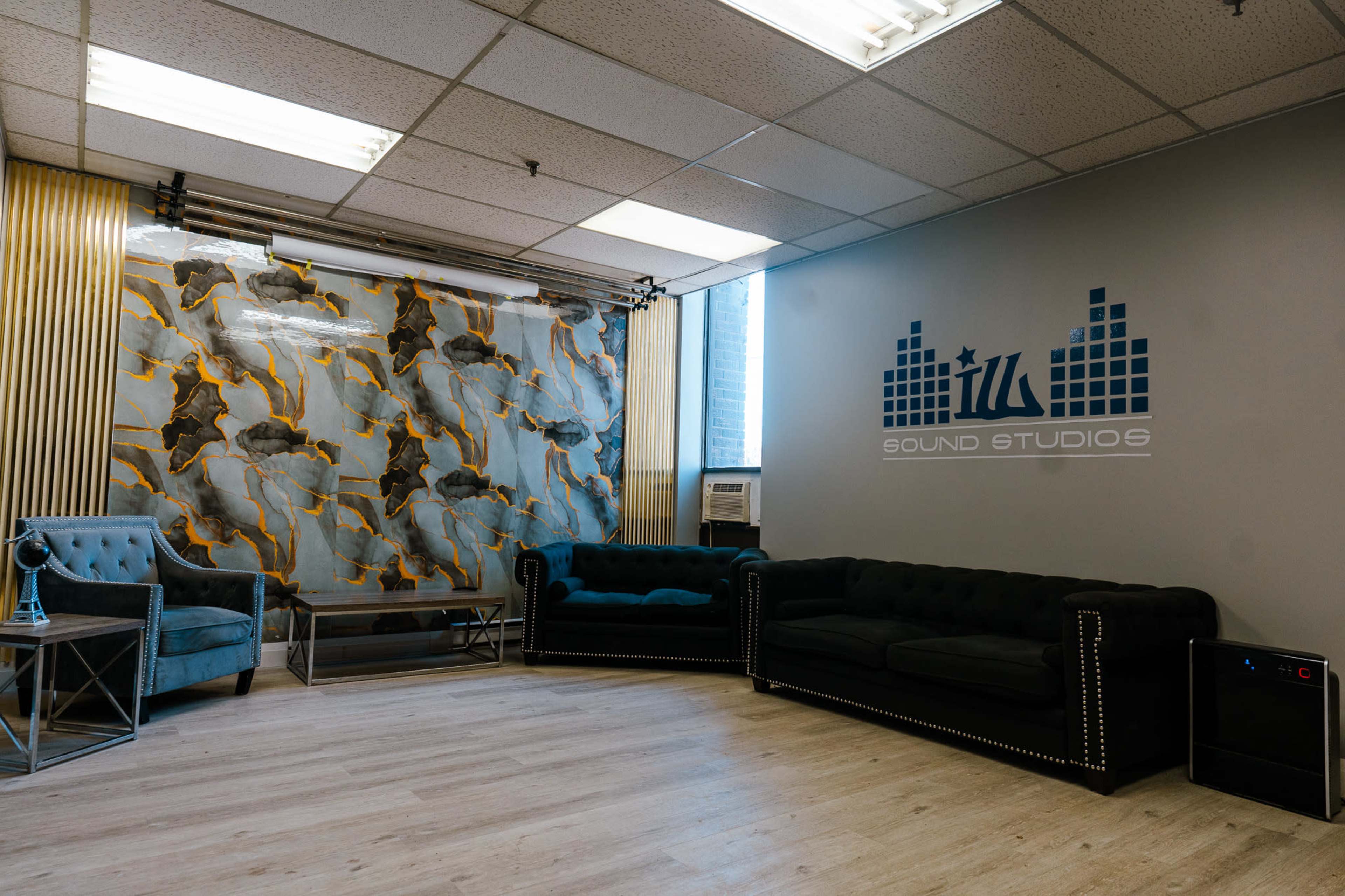 The image shows a modern studio lounge with two dark sofas, a glass coffee table, and a patterned backdrop featuring abstract designs on the wall.