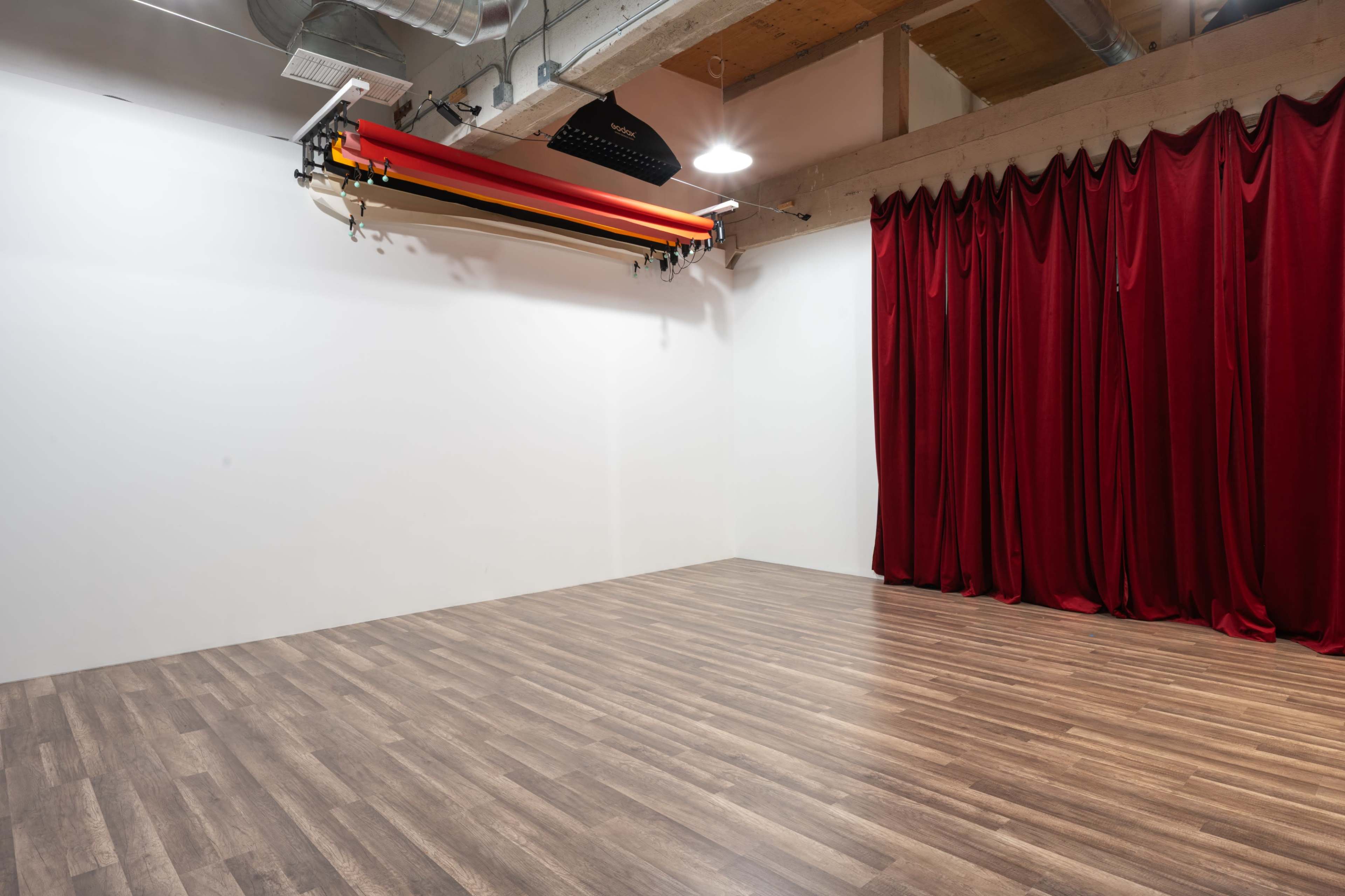 The image shows a studio space with wooden flooring, a white wall, and red curtains on one side.