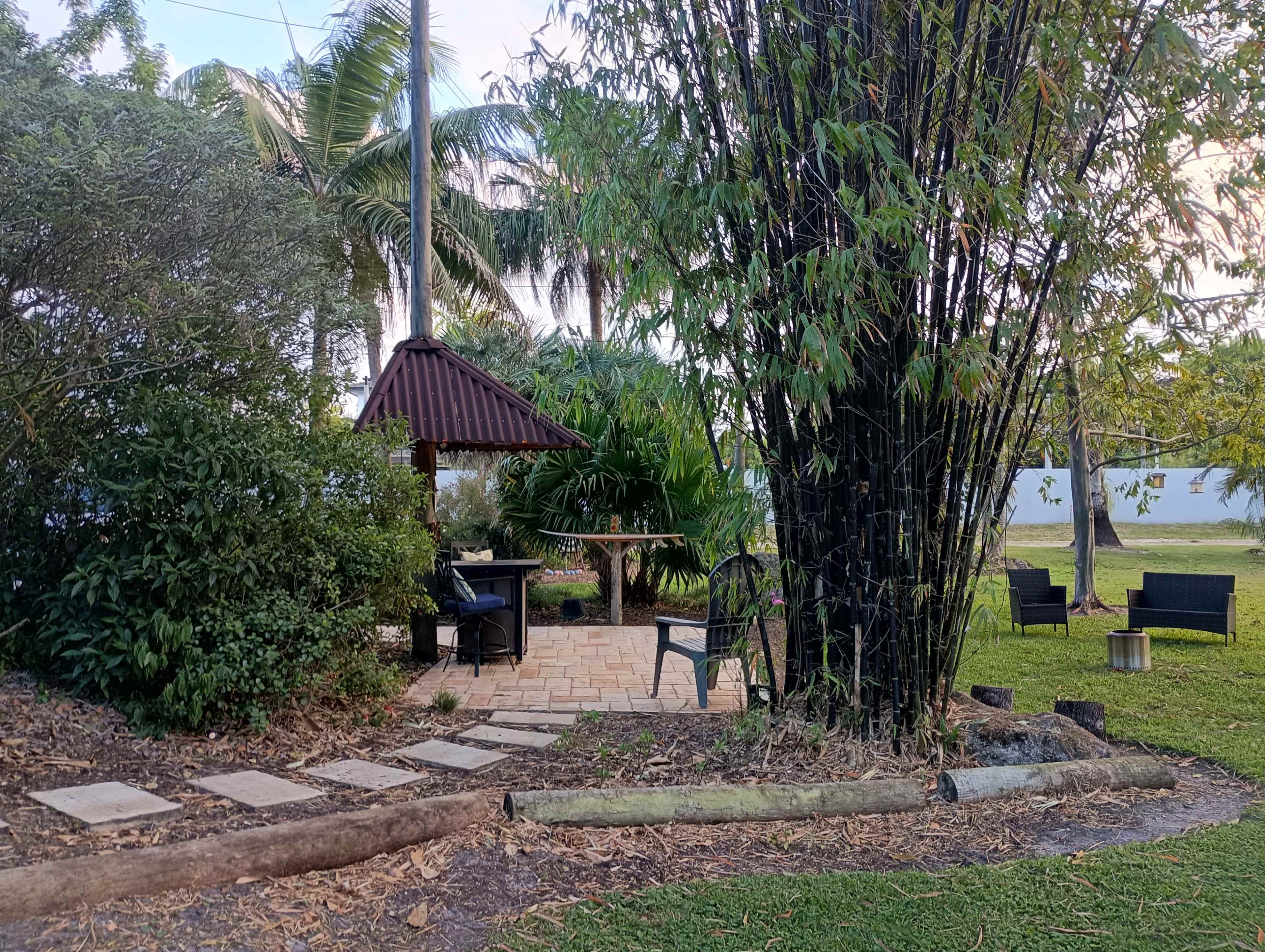 The image shows a landscaped garden area featuring a gazebo, chairs, a paved pathway, and bamboo plants, with tropical foliage surrounding the space.