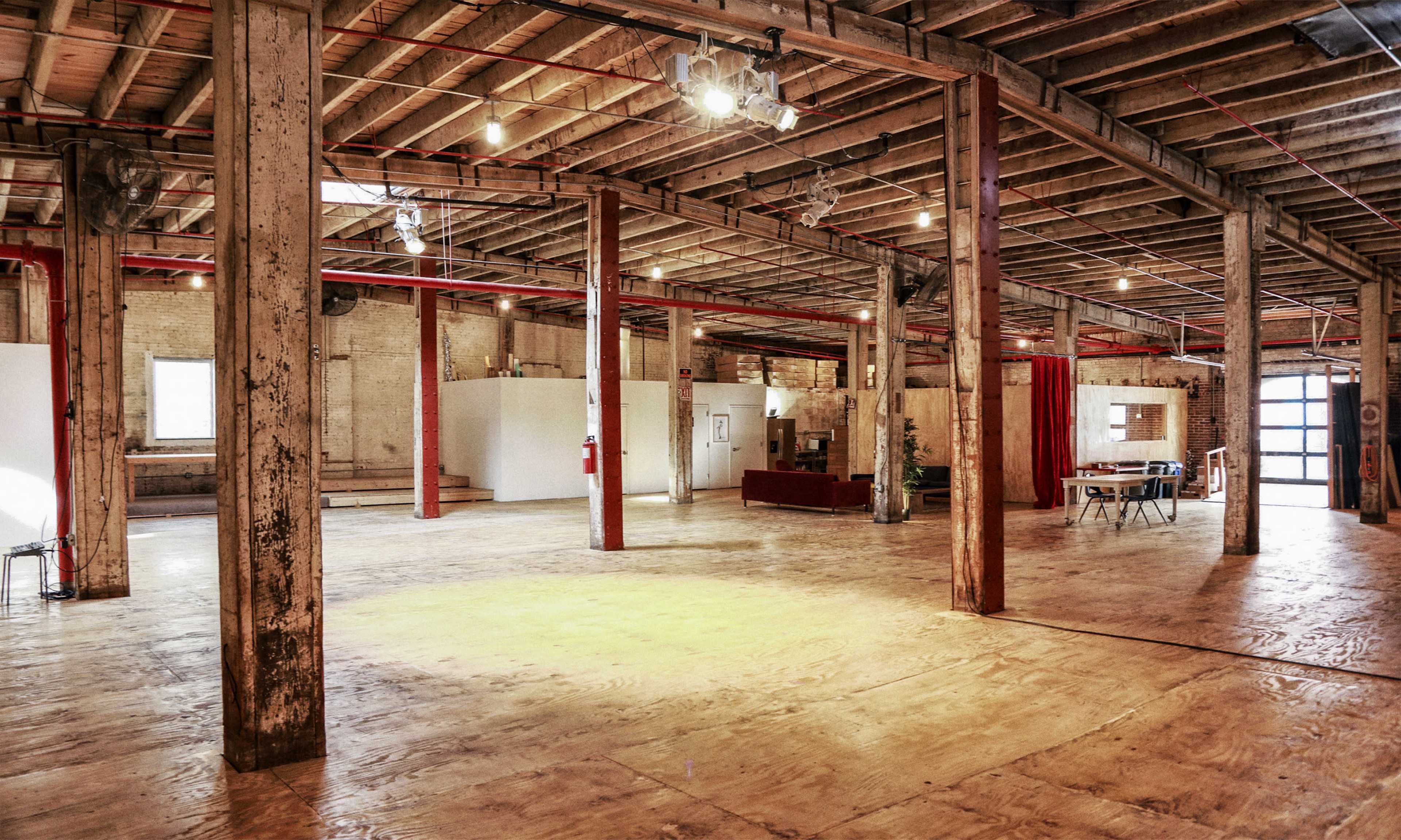 The image shows an expansive, empty industrial space with wooden beams, polished concrete flooring, and areas designated for seating and tables.