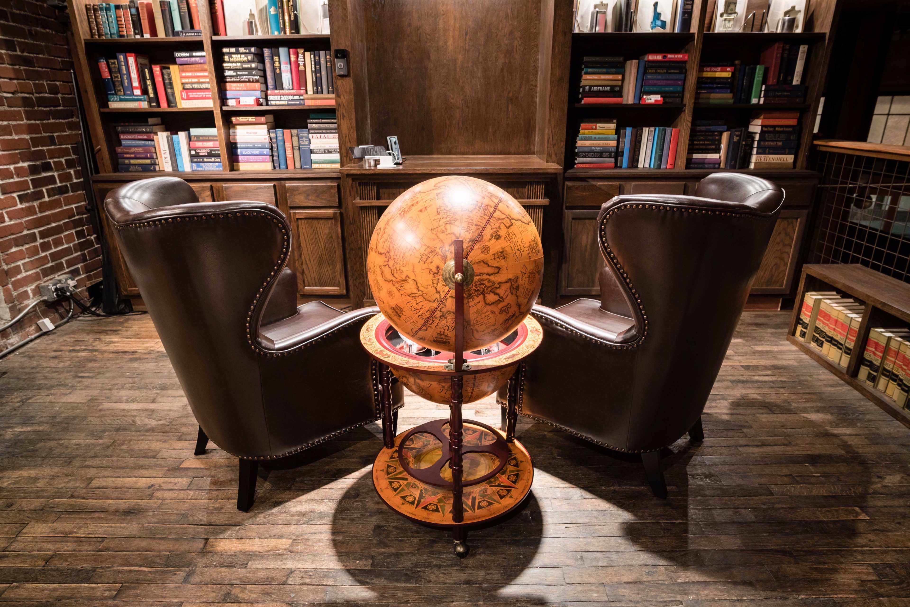 Two leather armchairs face a wooden globe on a stand, positioned between them in a room with bookshelves filled with books.