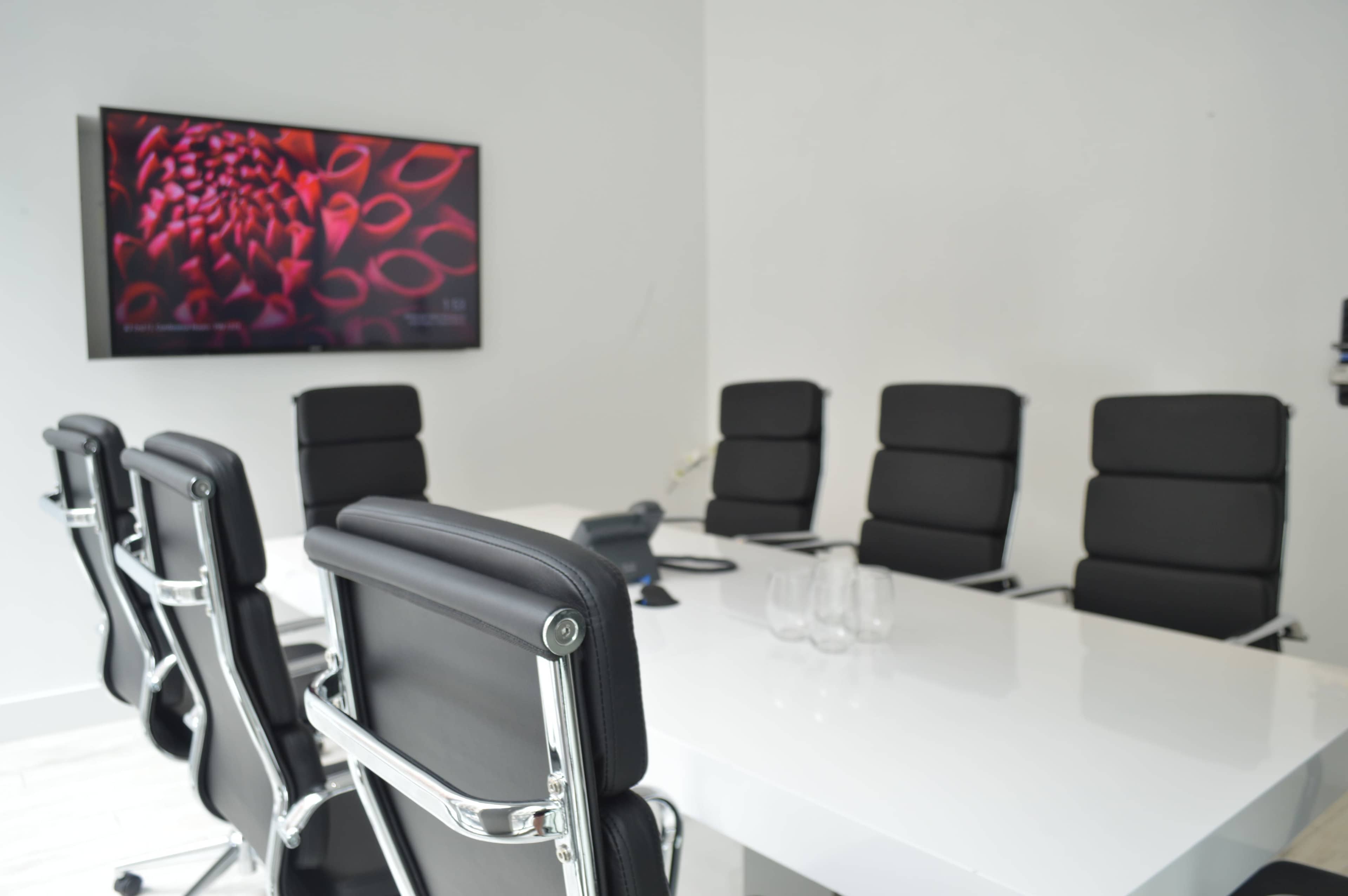 A modern conference room features a large white table surrounded by black chairs and a wall-mounted screen displaying colorful artwork.