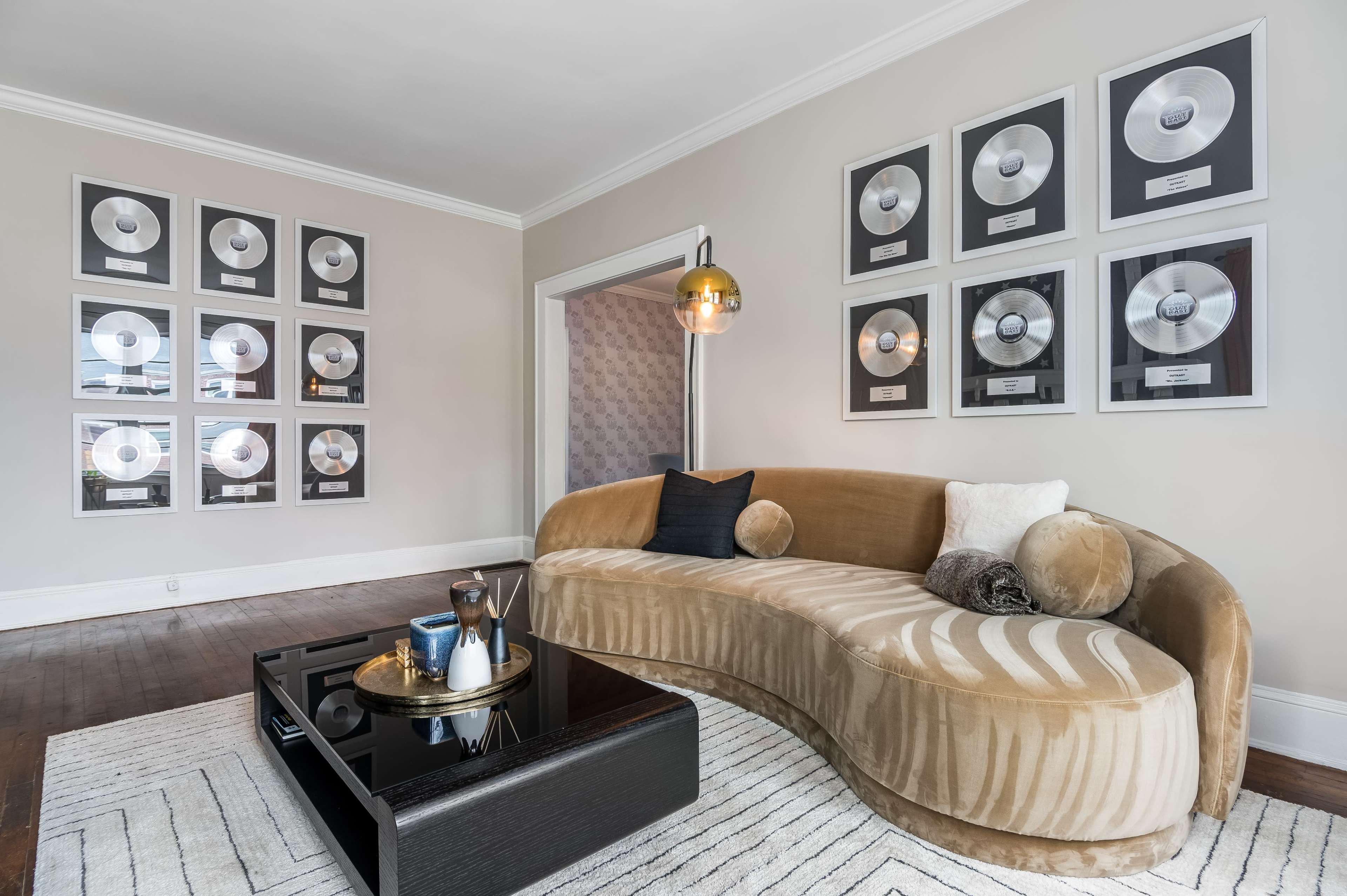 A curved, beige sofa is positioned in a living room adorned with silver record awards on the walls and a black coffee table in front.