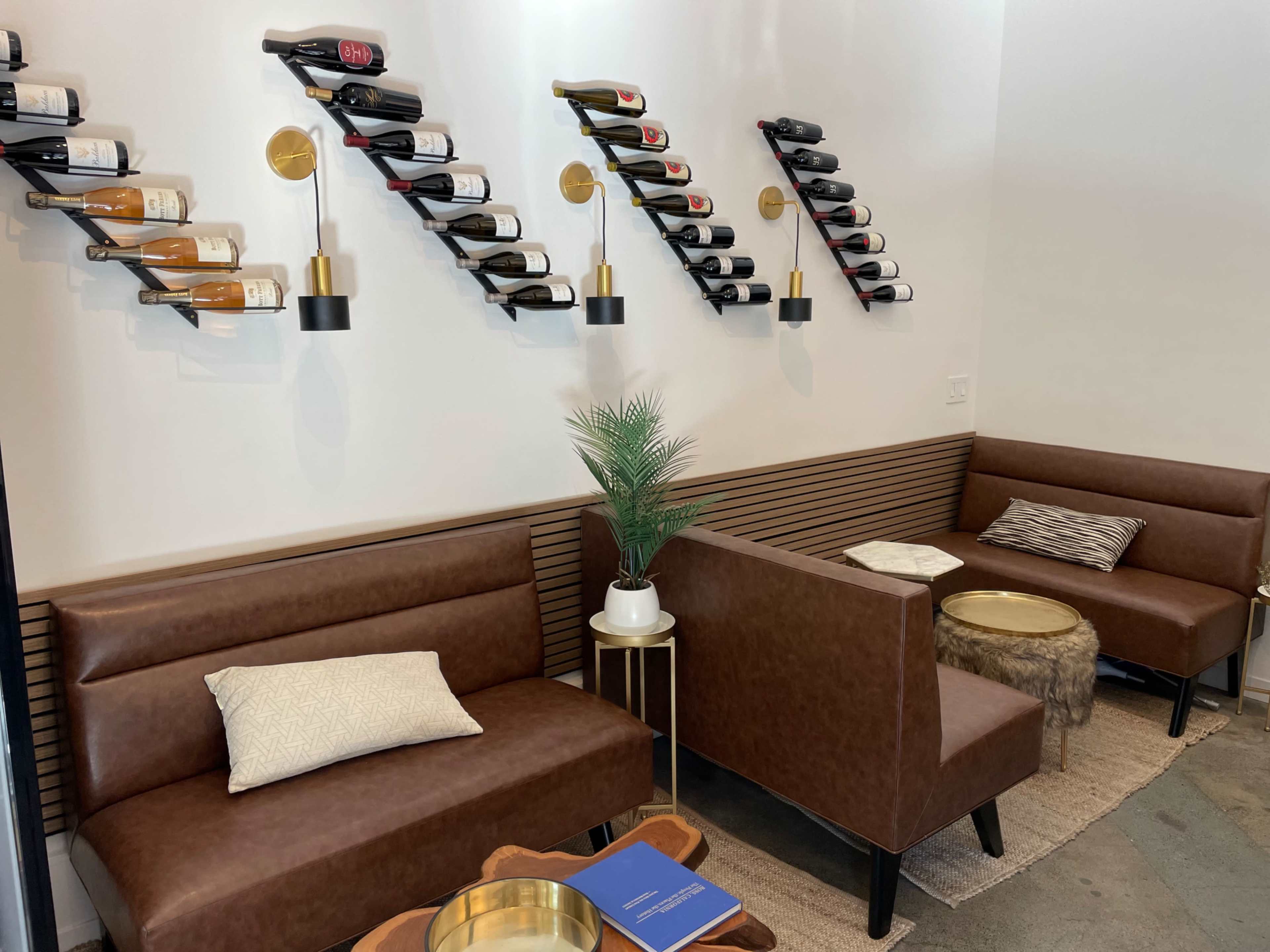 The image shows a modern seating area with brown couches, decorative wine racks on the wall, and a small table topped with a plant and a book on the floor.