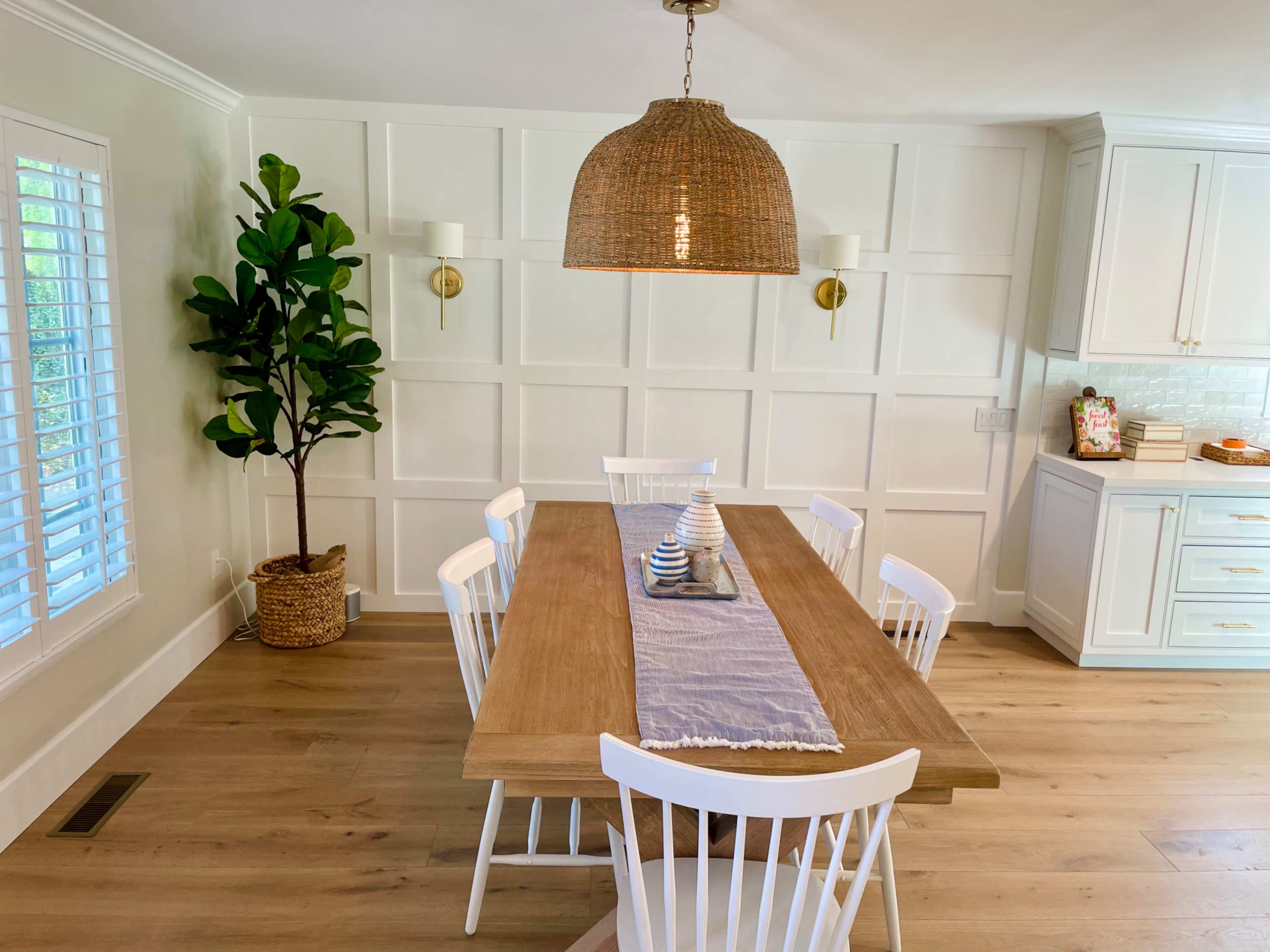 A dining area features a wooden table with white chairs, a center runner, and a large woven pendant light, alongside a leafy plant in a basket against a paneled wall.