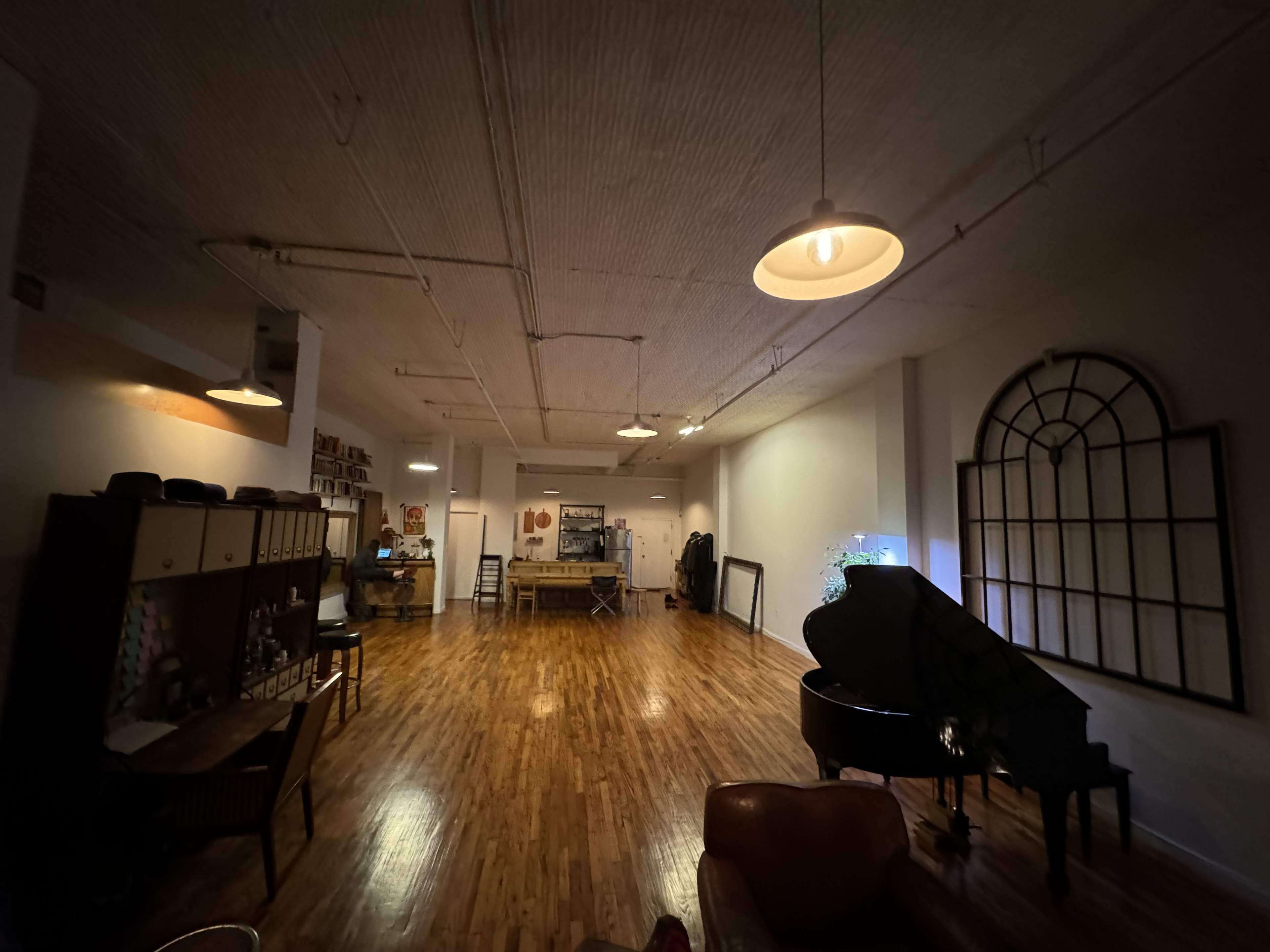 The image shows a spacious, well-lit room featuring a grand piano, wooden flooring, and various furniture pieces along the walls.