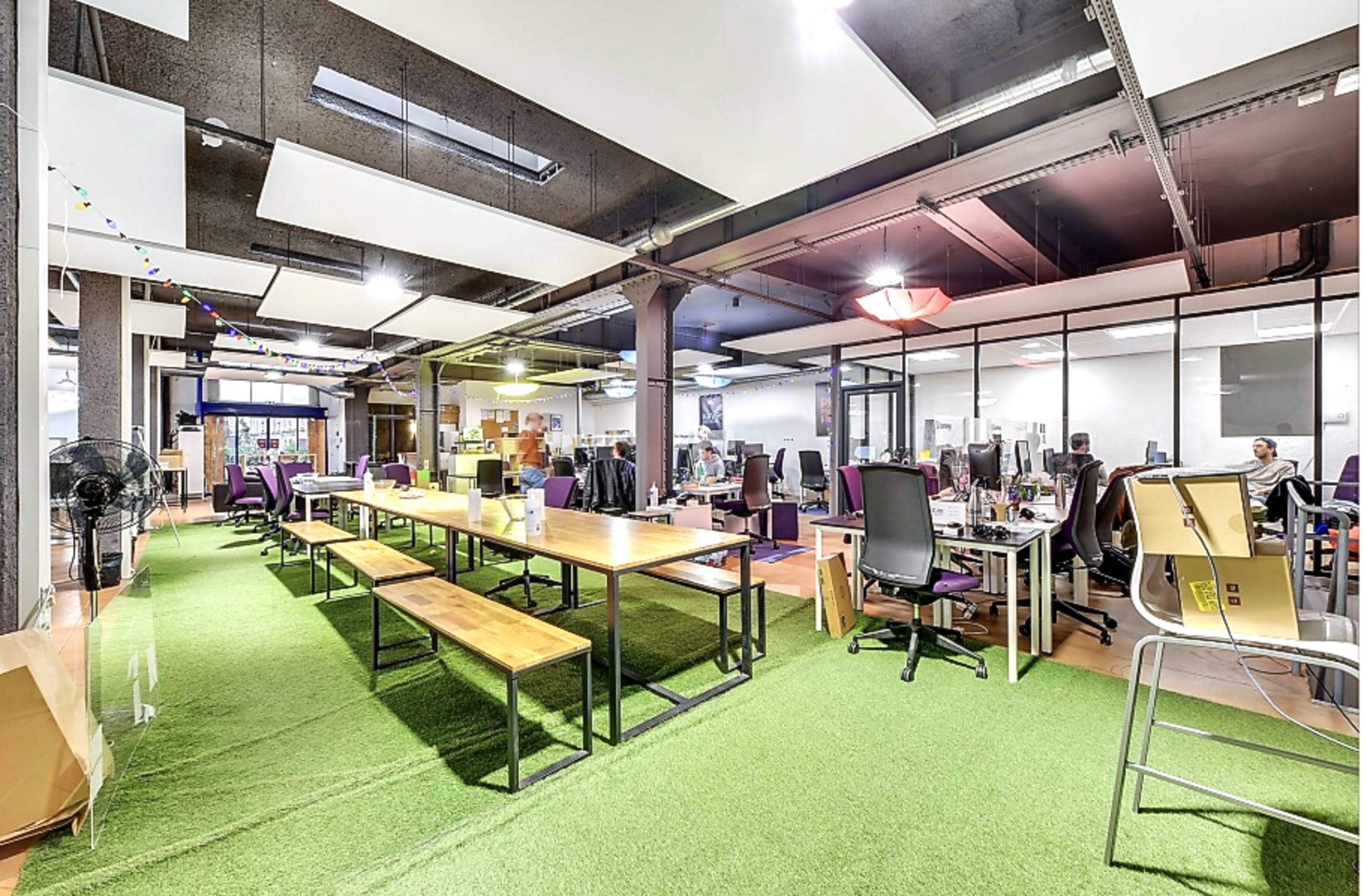 The image shows an open office space with long tables, chairs, and computer workstations, and artificial grass covering the floor.