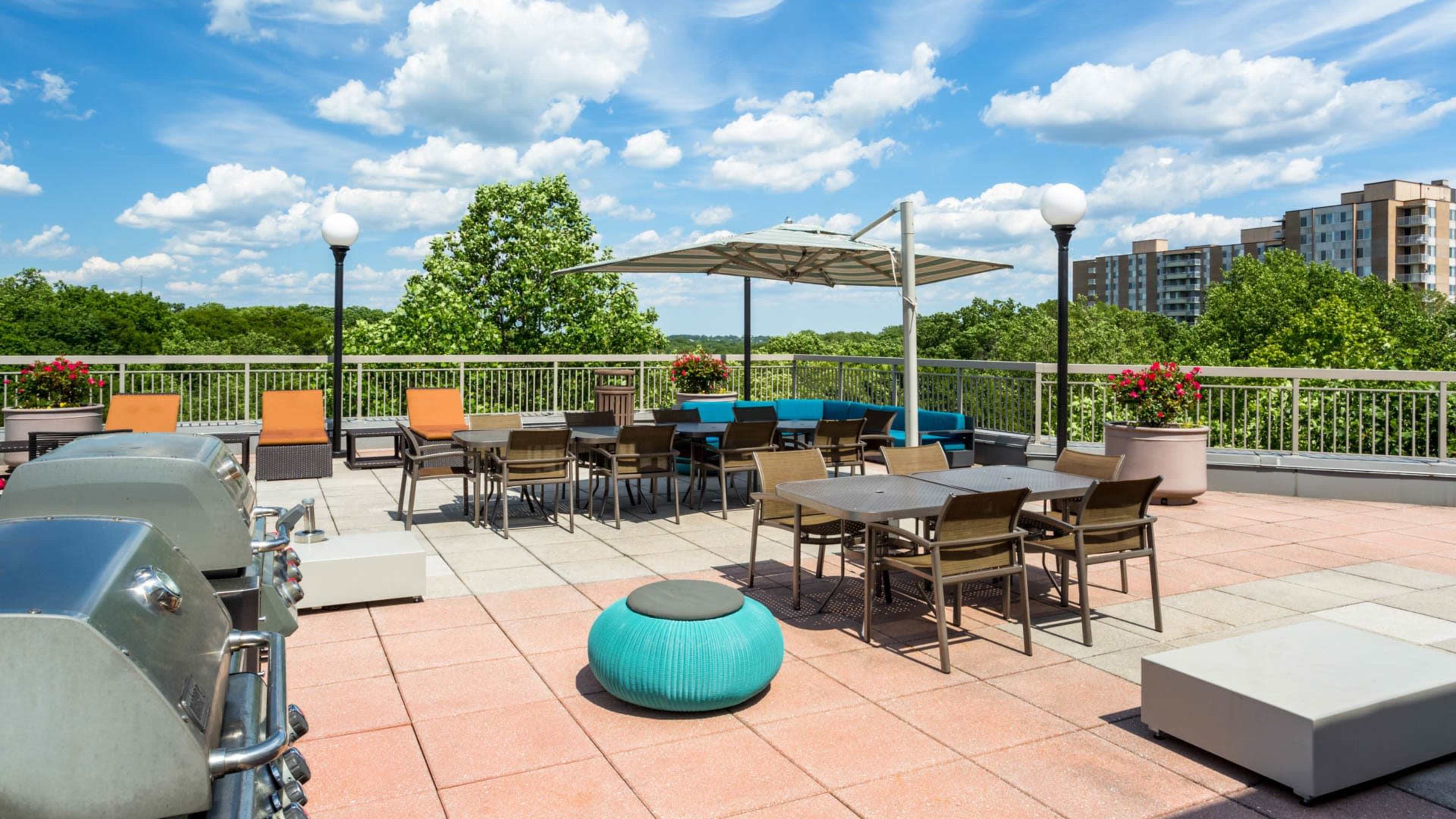 A rooftop patio area with seating, a barbecue grill, and a shaded table surrounded by greenery and apartment buildings in the background.