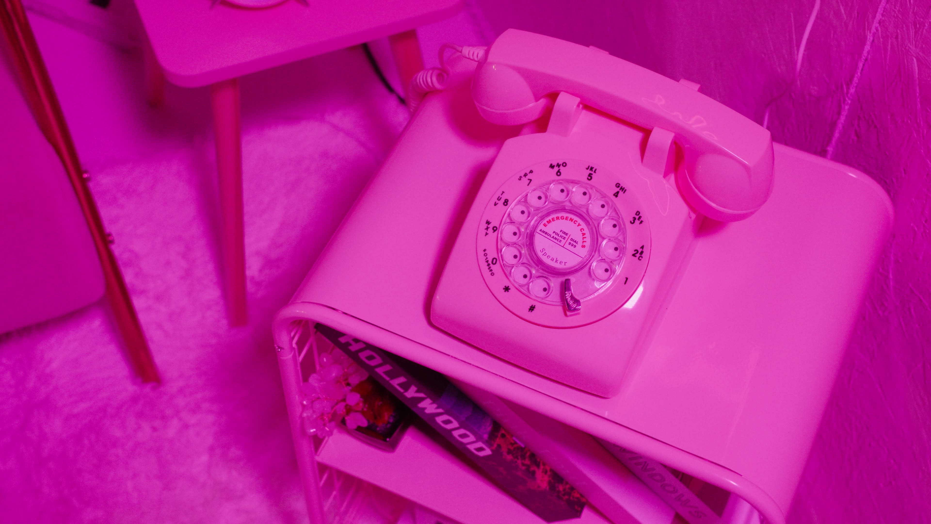A pink rotary telephone sits atop a pink table next to a shelf of books.