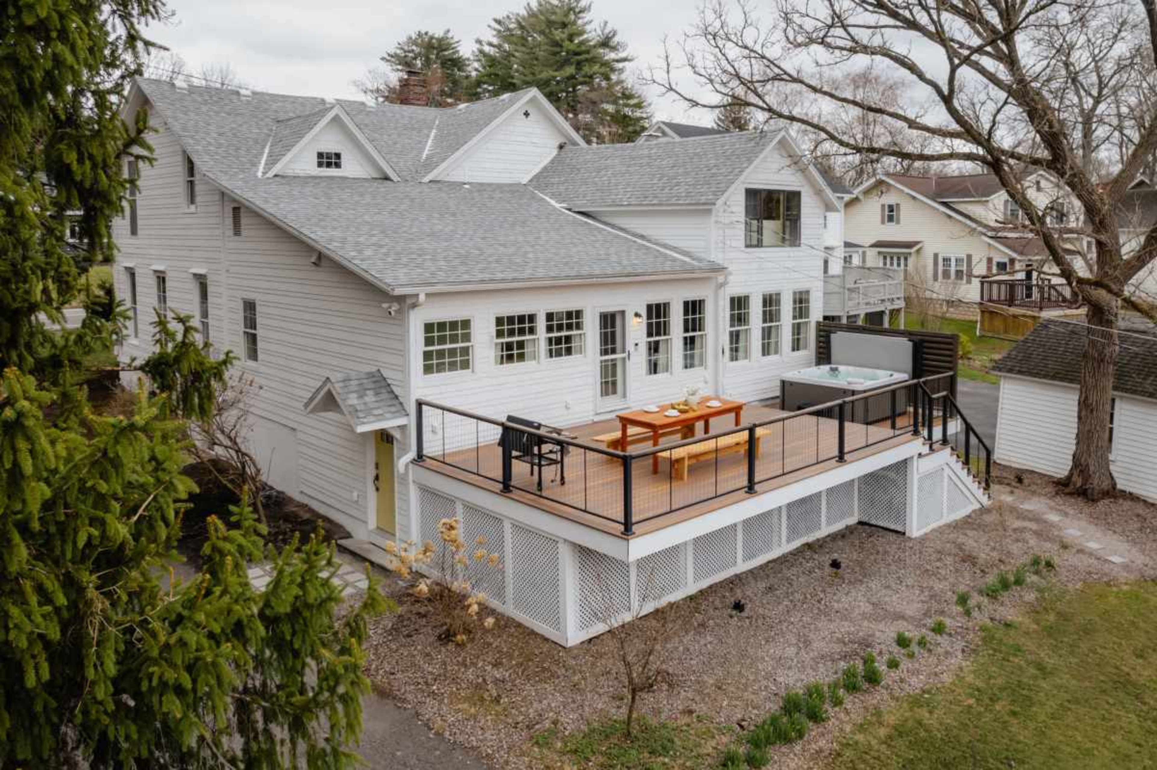 The image shows a two-story white house with a large wooden deck and outdoor seating area, surrounded by trees and other residential buildings.