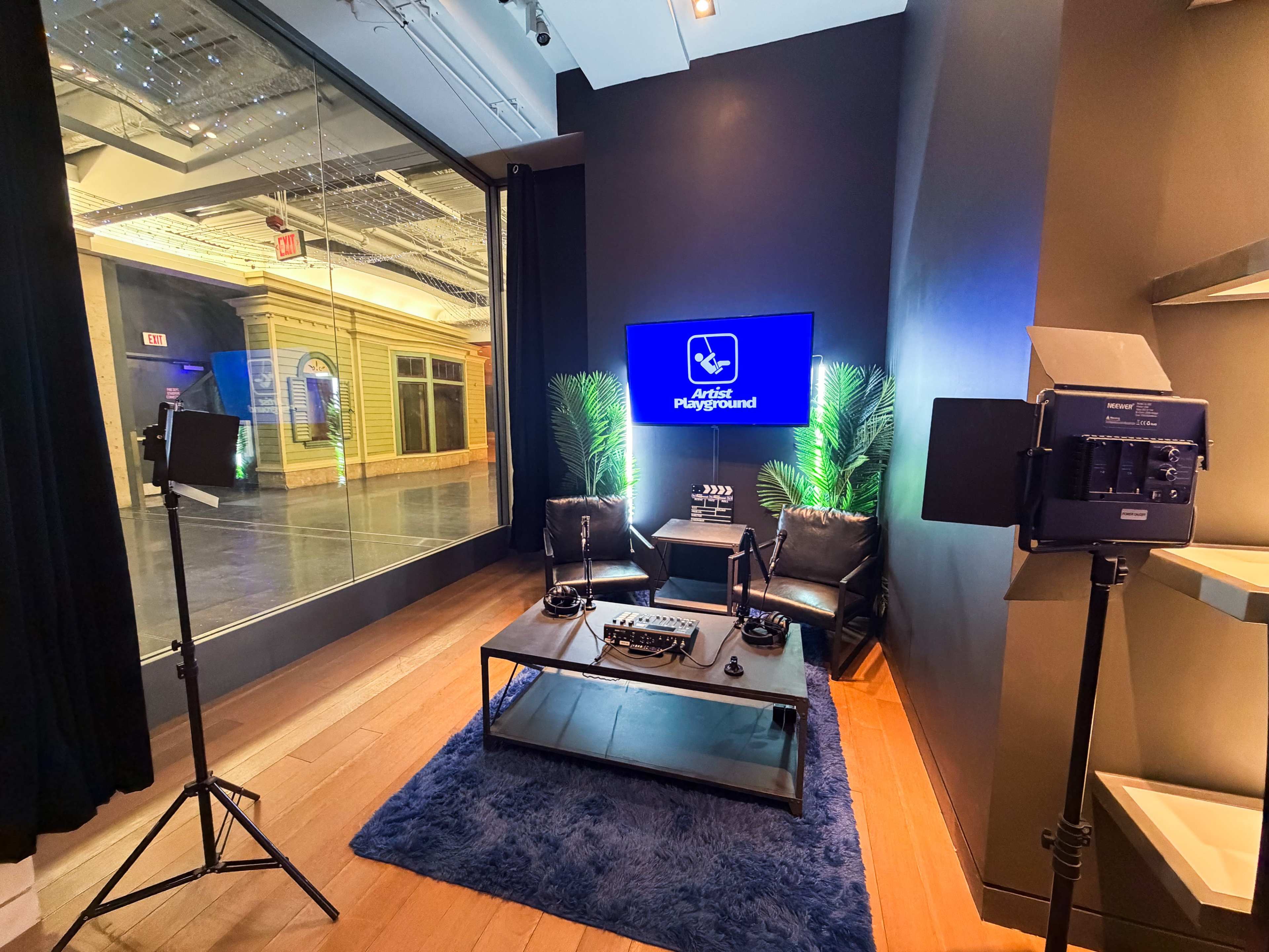 The image shows a modern recording studio setup featuring two chairs, a blue rug, and professional lighting equipment, along with a screen displaying a logo.