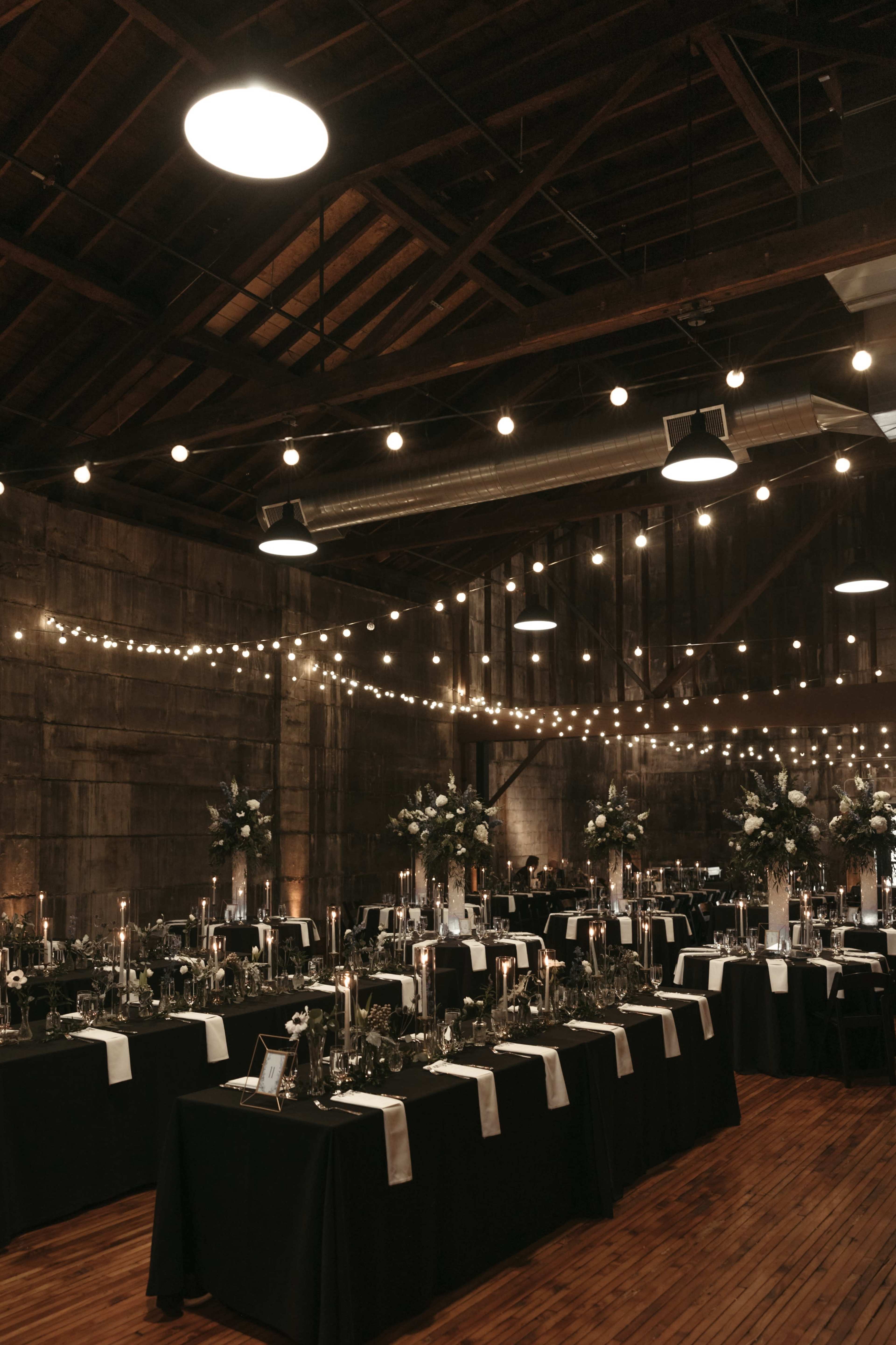 A spacious indoor venue is set with multiple tables covered in black cloth, adorned with candles and floral centerpieces, and illuminated by string lights overhead.